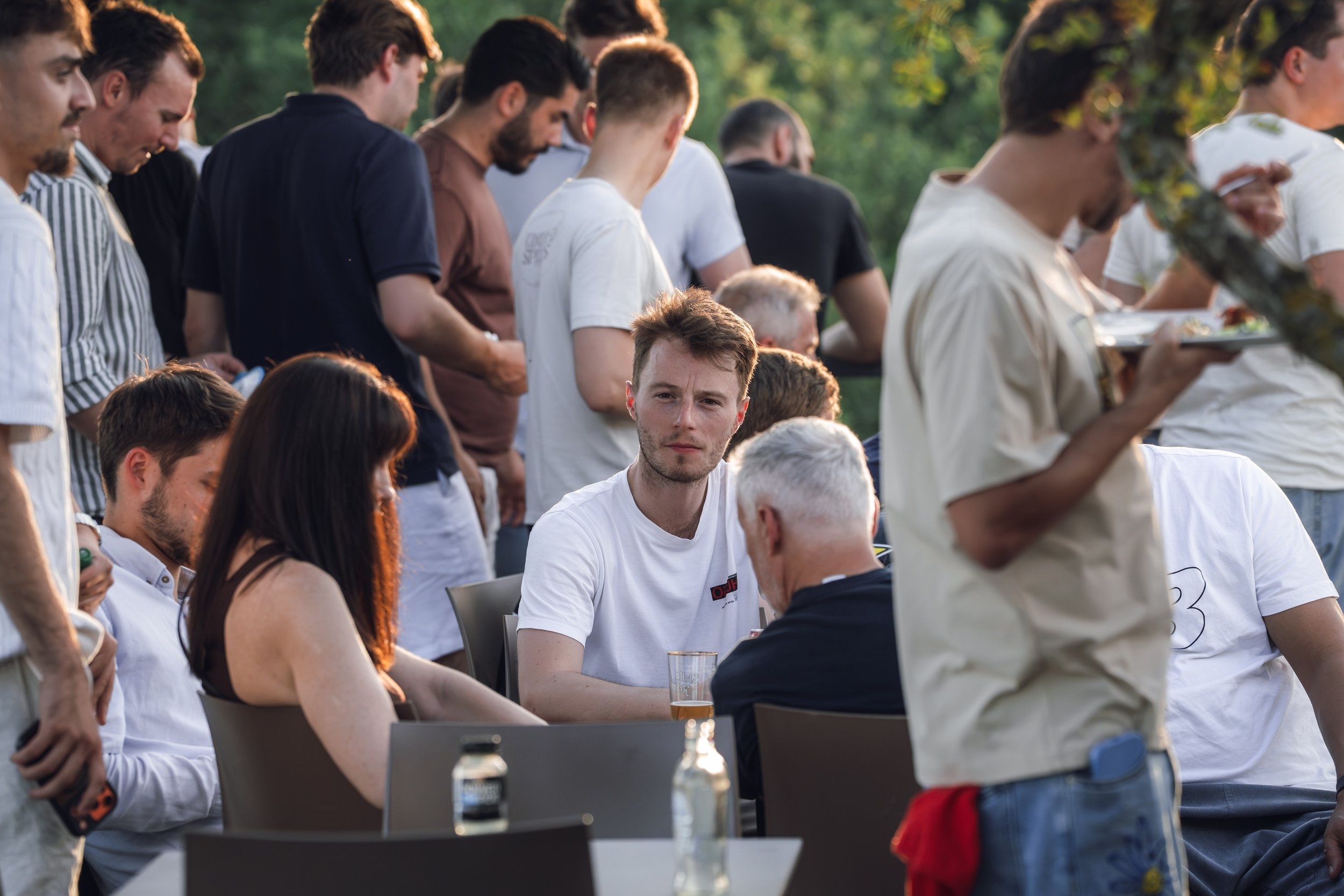 A group of people gathered outdoors, some standing and some sitting, during a social event or gathering in the evening, with drinks on the tables and blurred green trees in the background.
