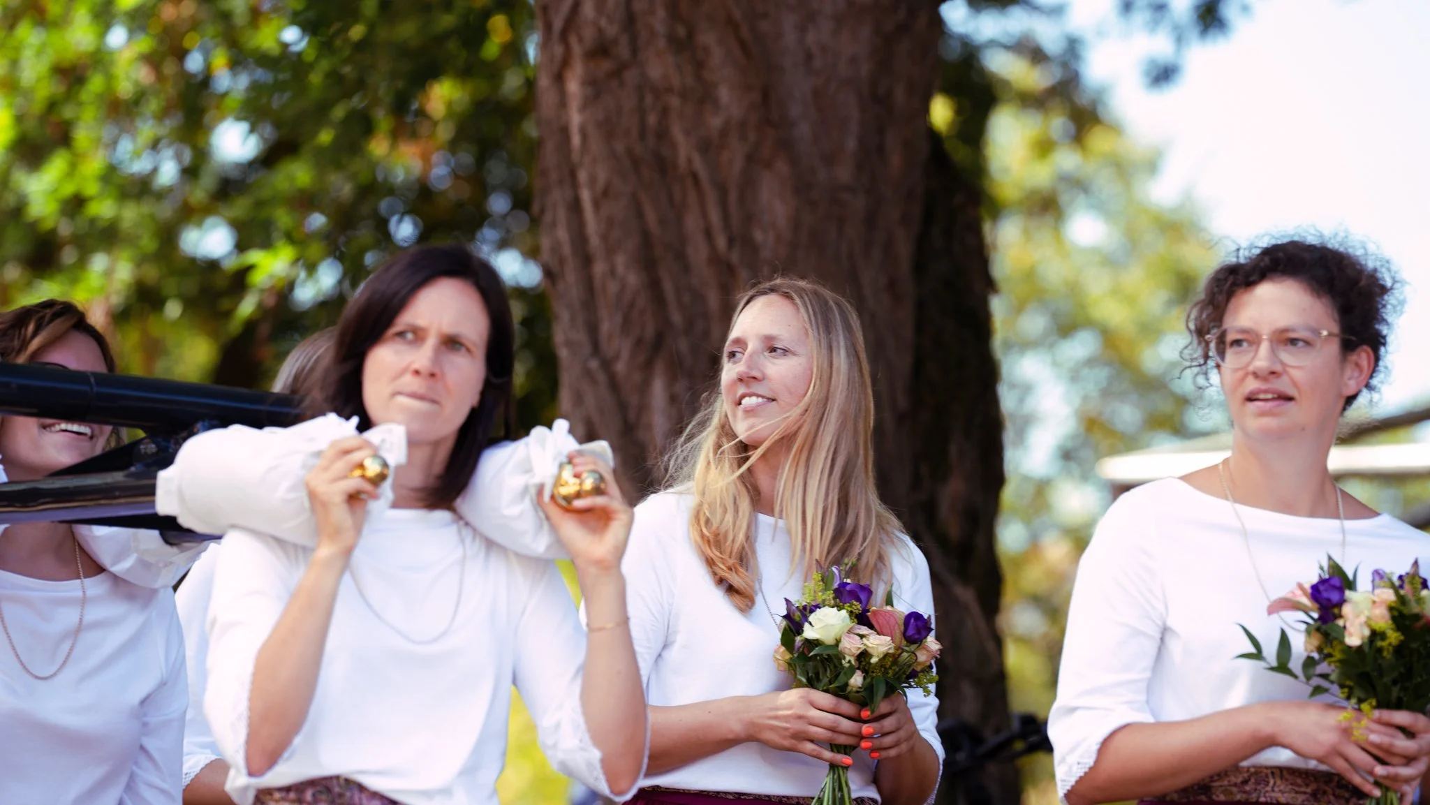 Group of women standing outdoors, one holding a bouquet of flowers, another holding a small gift box, and a third with their hands clasped, standing near a large tree with blurred green leaves in the background.