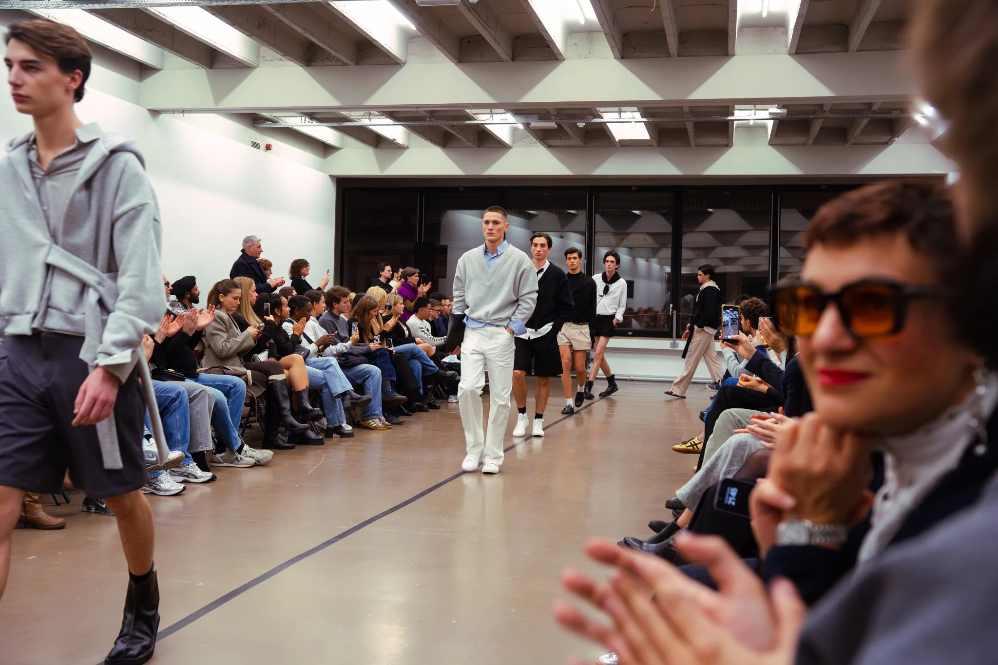 Models walking on a runway during a fashion show with an audience seated on both sides. The audience is clapping and taking photos.