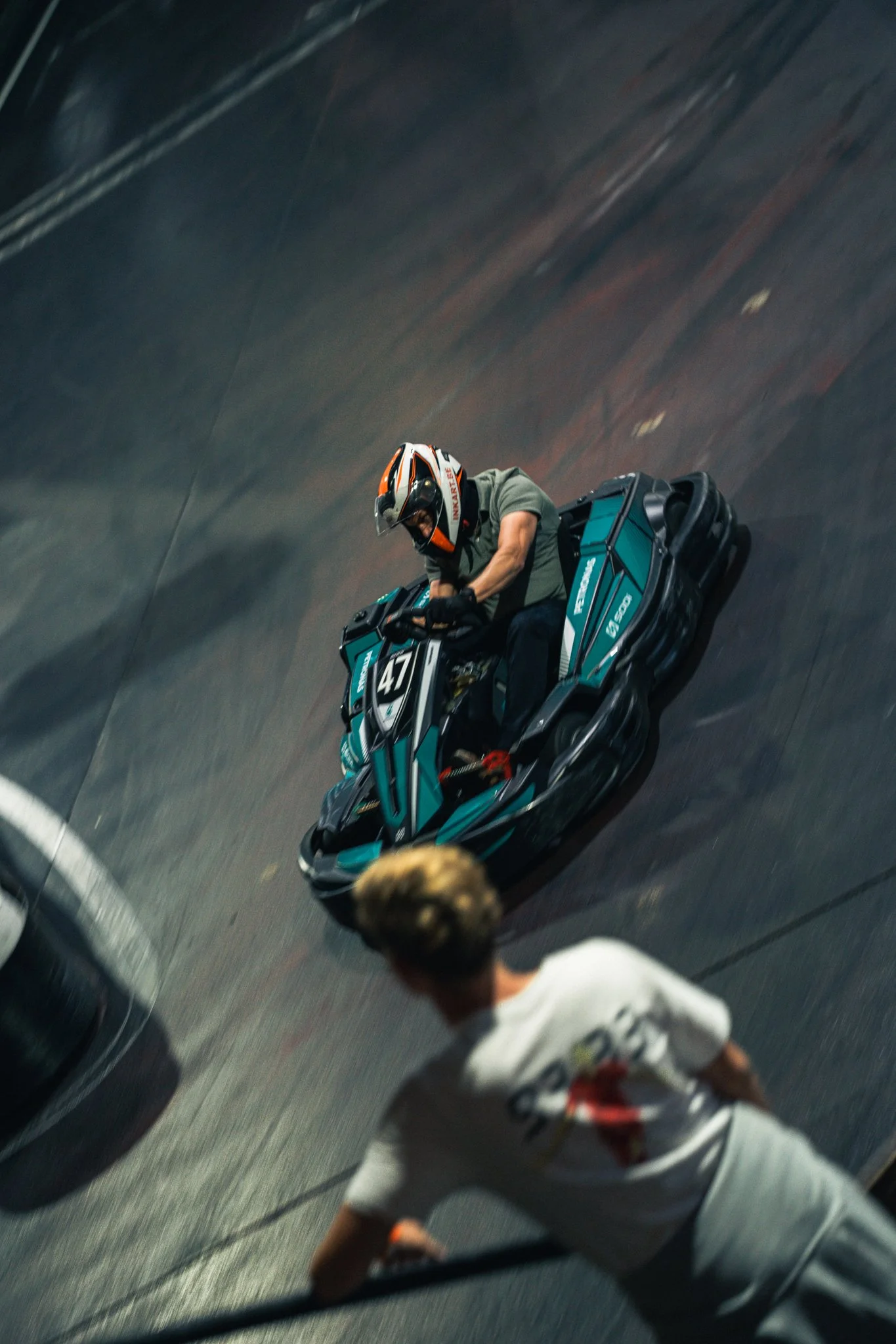 A person riding a teal and black bumper car on an indoor track, wearing a helmet and casual clothes. Another individual with blonde hair wearing a white shirt and gray pants is seen in the foreground, possibly supervising or observing.