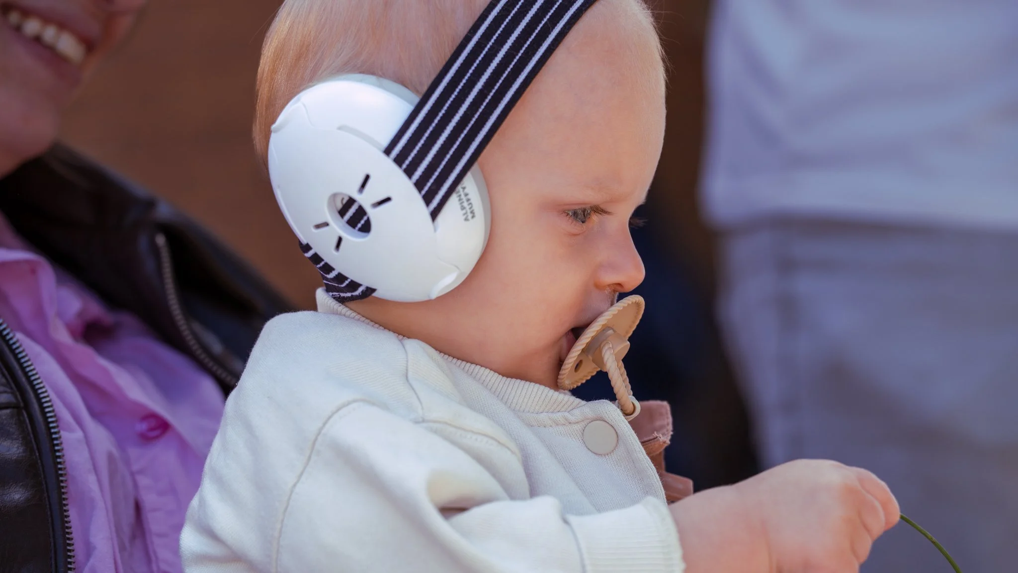 Young boy with blond hair wearing large white headphones with black stripes, holding a green stem and a pacifier in his mouth.