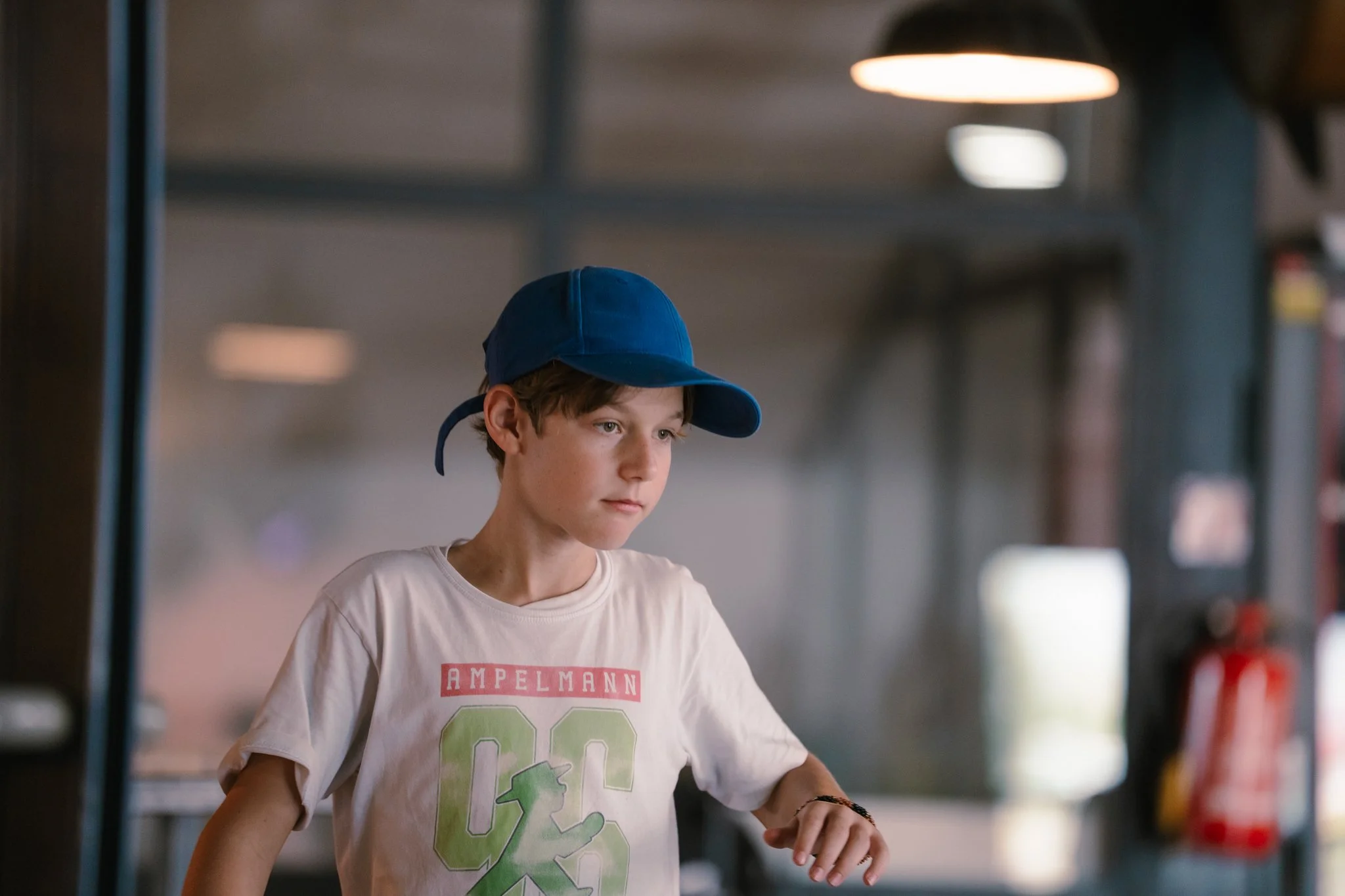 A young boy wearing a blue baseball cap and a white T-shirt with a graphic design, looking down with a focused expression, in an indoor setting with industrial lighting and a blurred background.