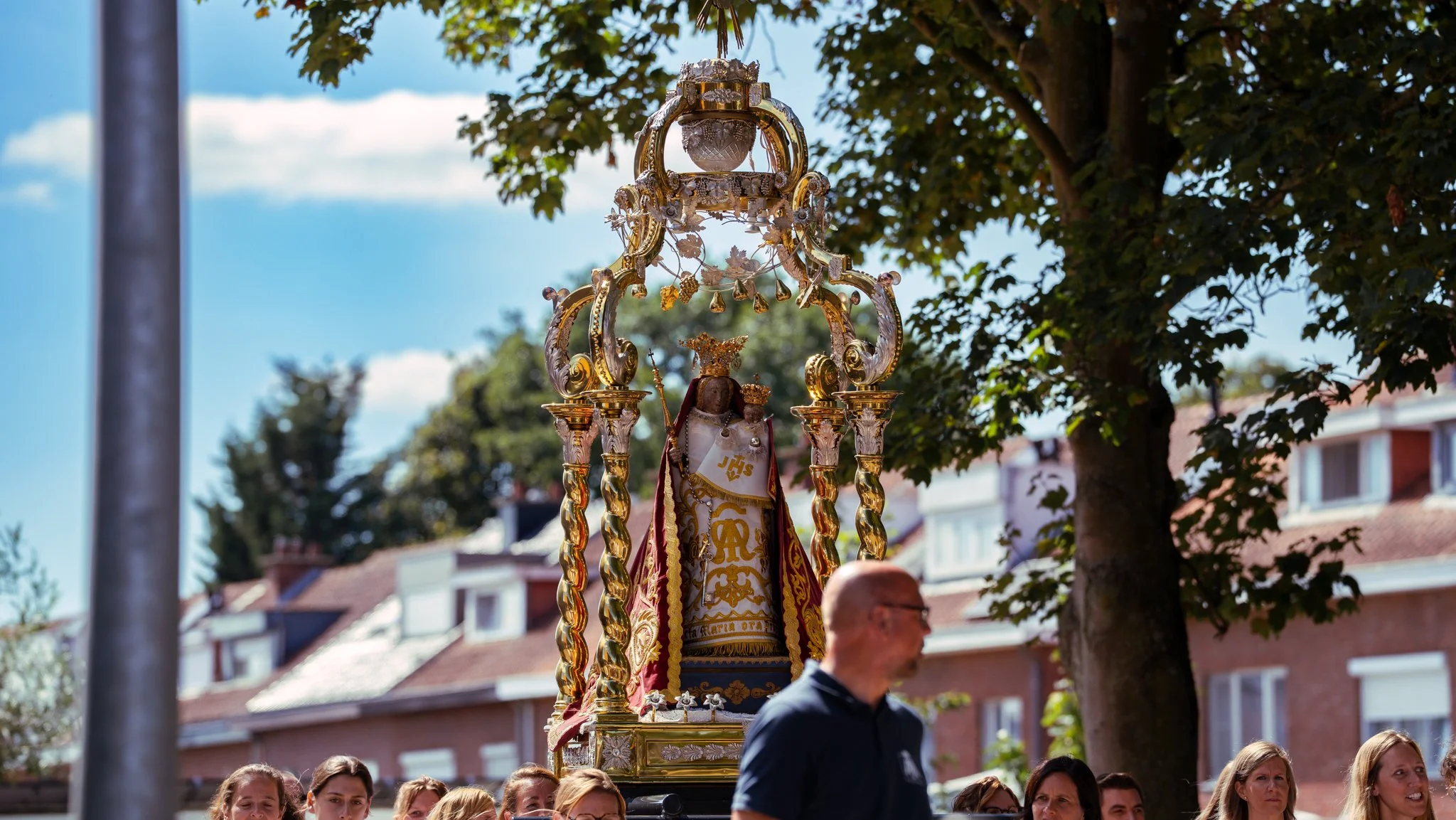 A religious procession featuring a large ornate statue of the Virgin Mary with a crown, robes, and religious symbols, carried through a neighborhood street with spectators and a man walking past.