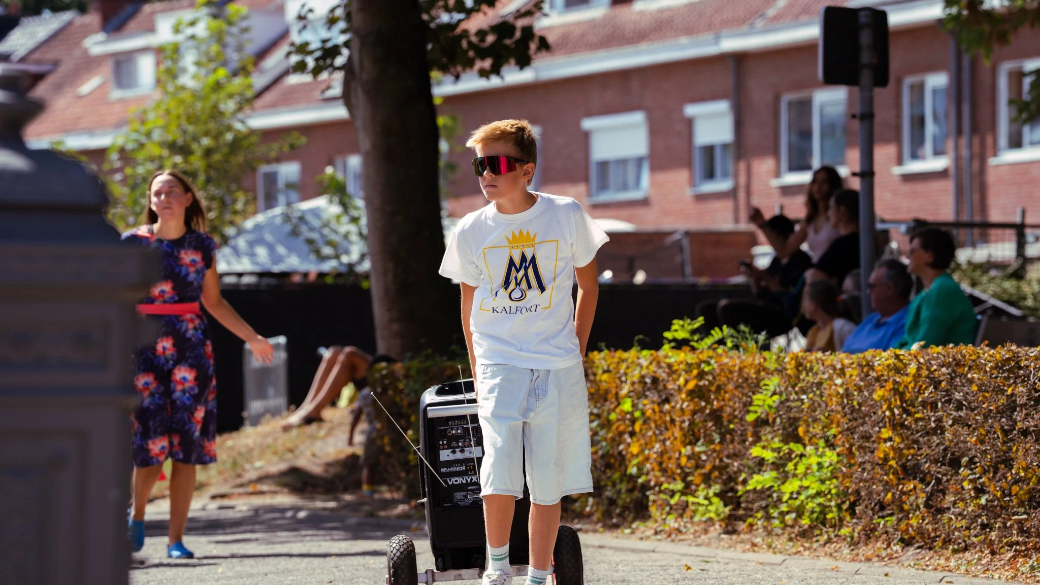 A young boy wearing sunglasses, a white t-shirt with a logo, and shorts, standing outdoors on a sidewalk, with a small electric skateboard or scooter adjusted with wires, beside him. In the background, a woman in a floral dress and several people sit