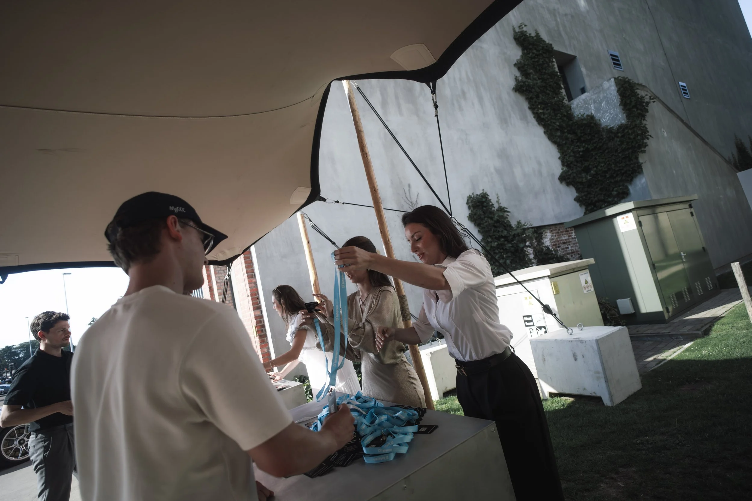 People gather under a canopy to receive name tags and registration materials at an outdoor event, with a concrete building, greenery, and utility boxes visible in the background.