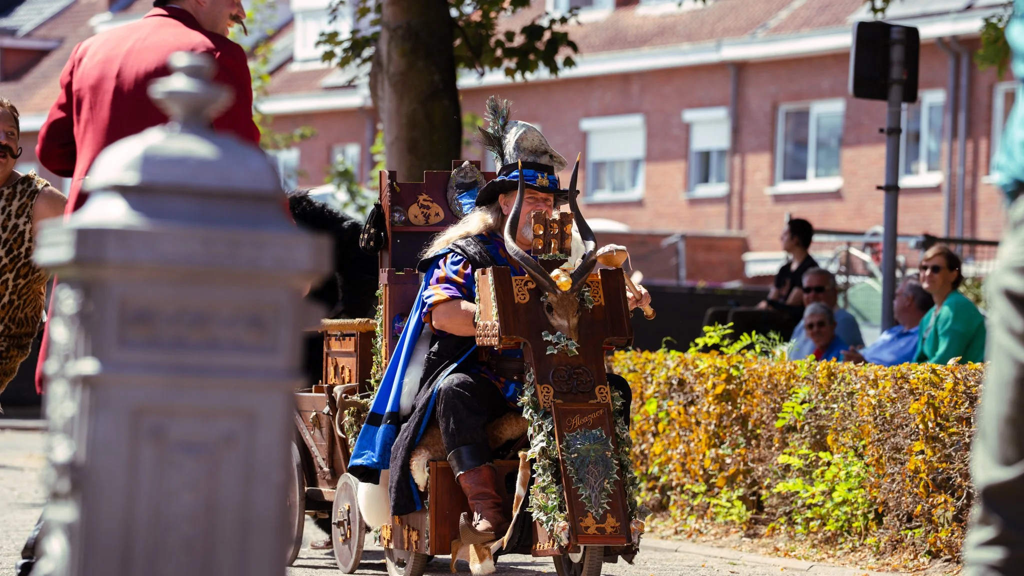 Person dressed as a wizard or medieval character sitting in a decorated cart during a parade, surrounded by onlookers and on a sunny day.