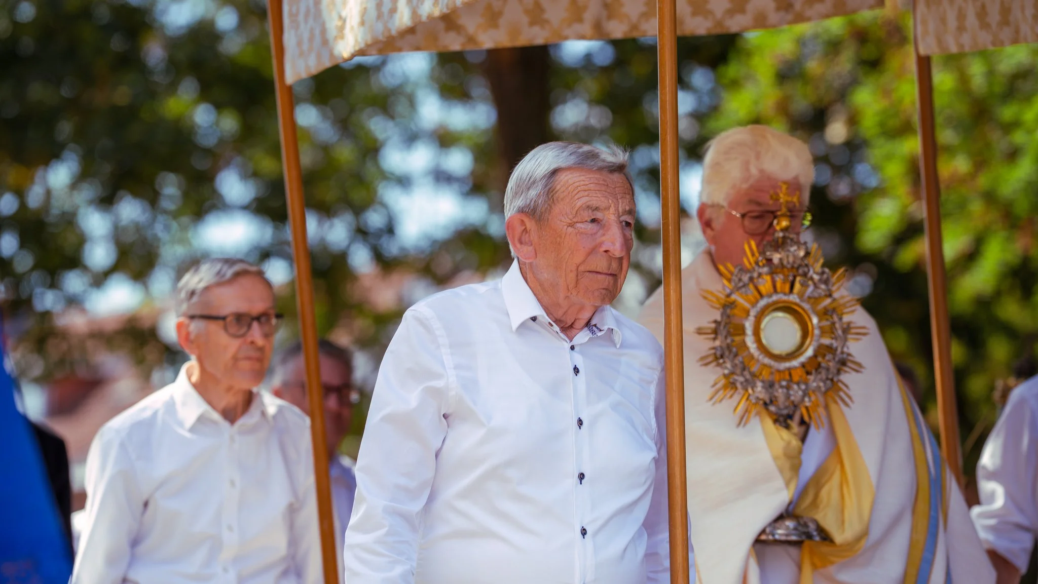 An elderly man in a white shirt participating in a religious procession, walking under a canopy, with other men nearby, one of whom is wearing ceremonial attire with a large ornate medal, all outdoors with trees in the background.
