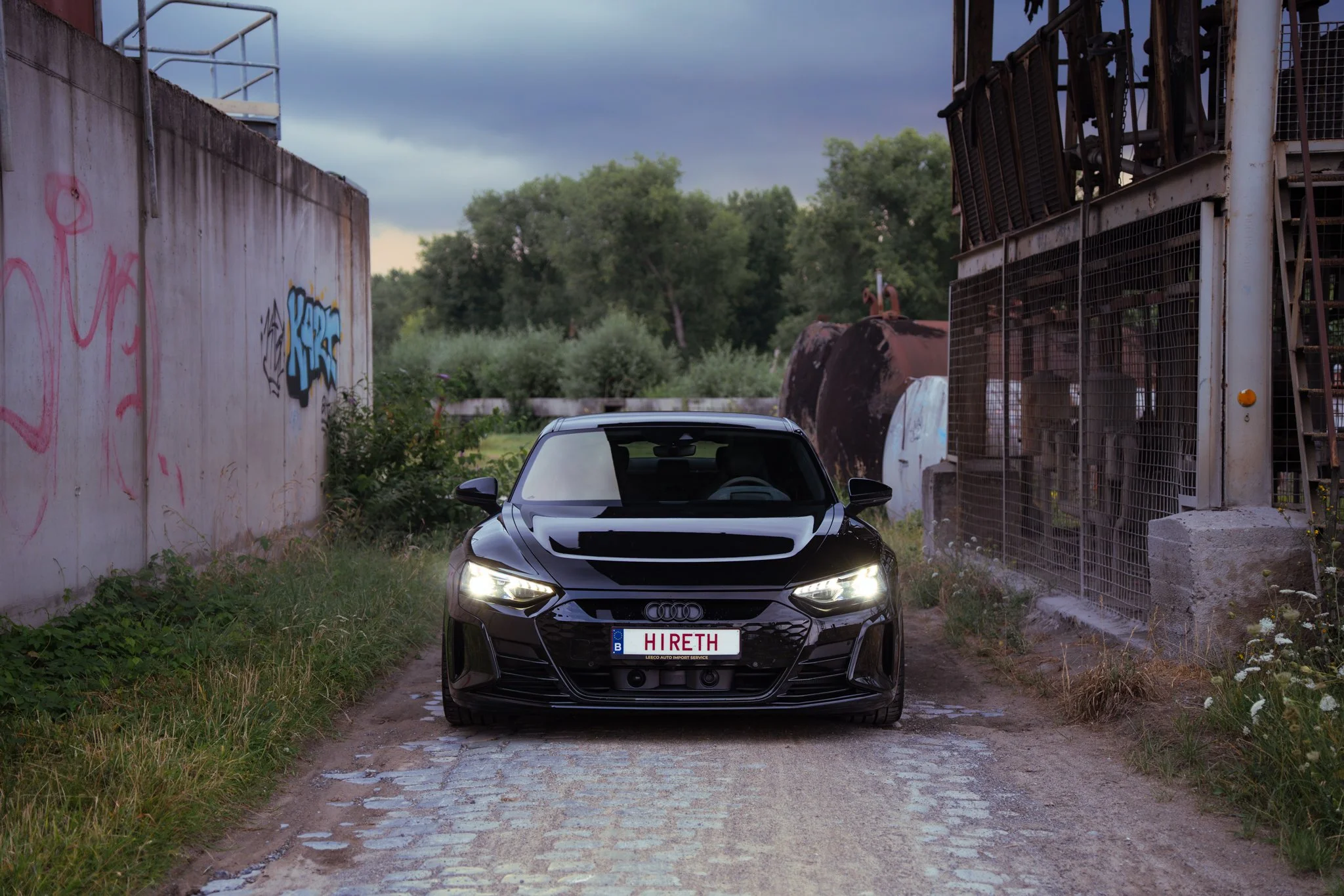 Black Audi car parked on a gravel path between graffiti-covered concrete wall and industrial structures with greenery in the background.