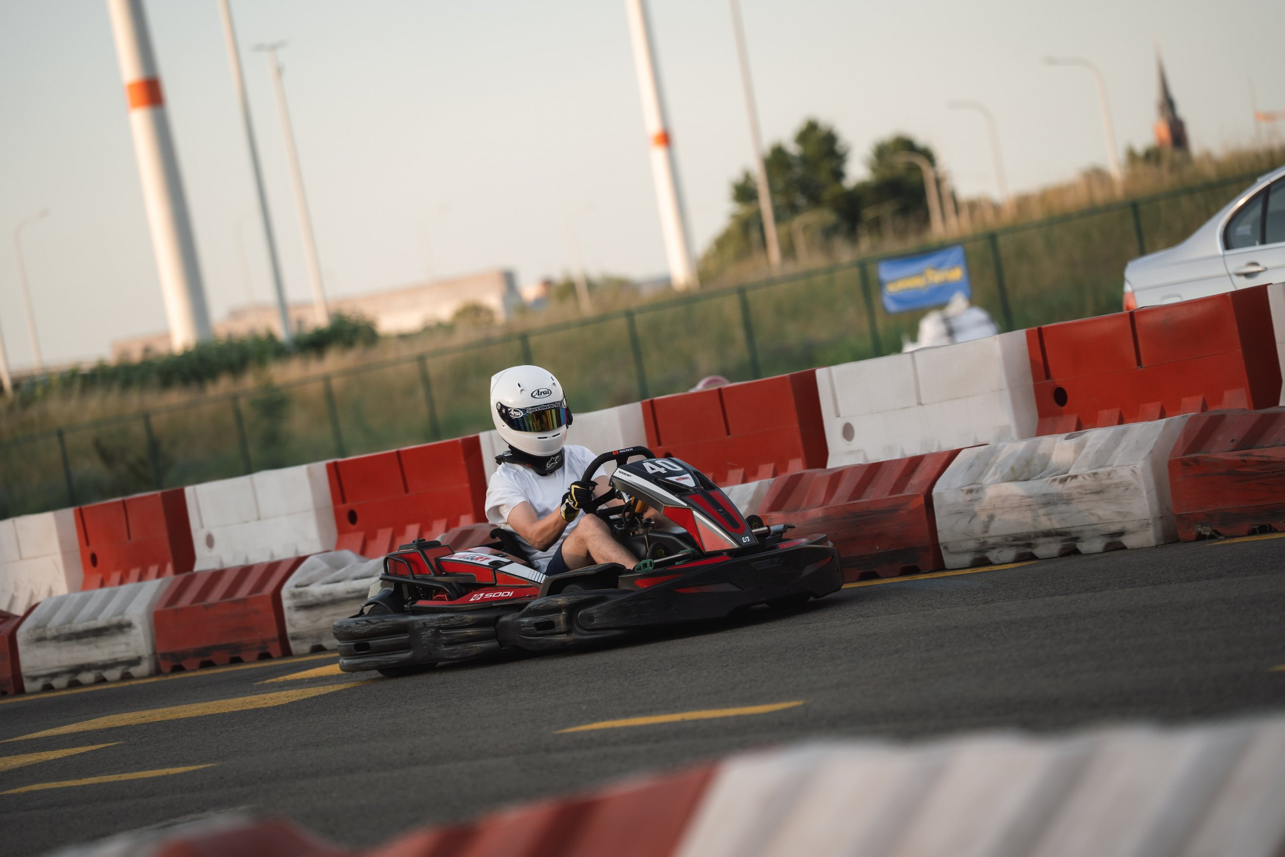 A person wearing a white helmet and white shirt driving a red and black go-kart on an outdoor track with orange and white barriers, with a background of trees, streetlights, and a distant church steeple.