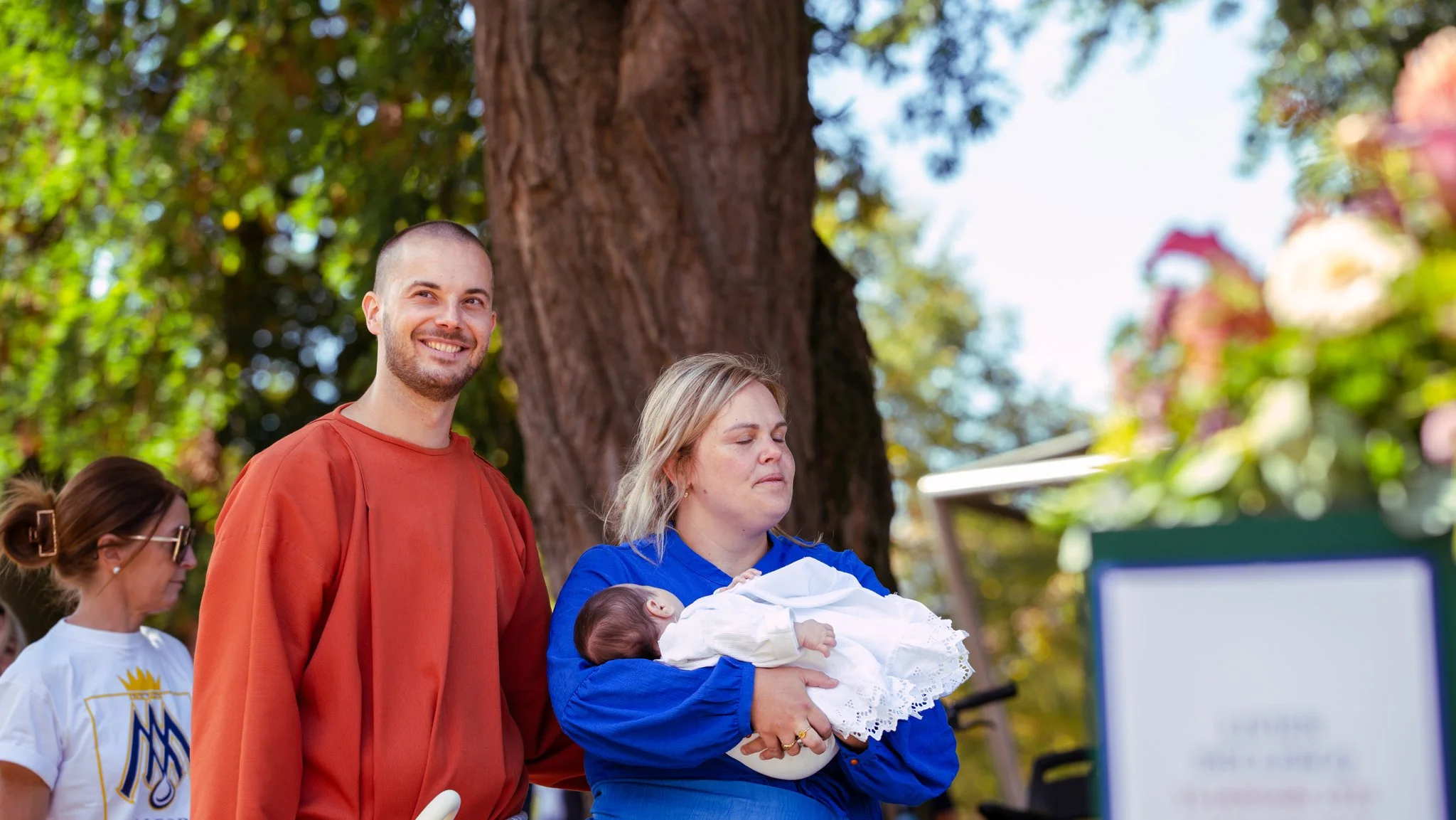 A woman with blonde hair holding a newborn baby dressed in white outdoors in front of a large tree. A man with a shaved head and beard, wearing an orange shirt, stands nearby smiling. Other people are partially visible in the background.