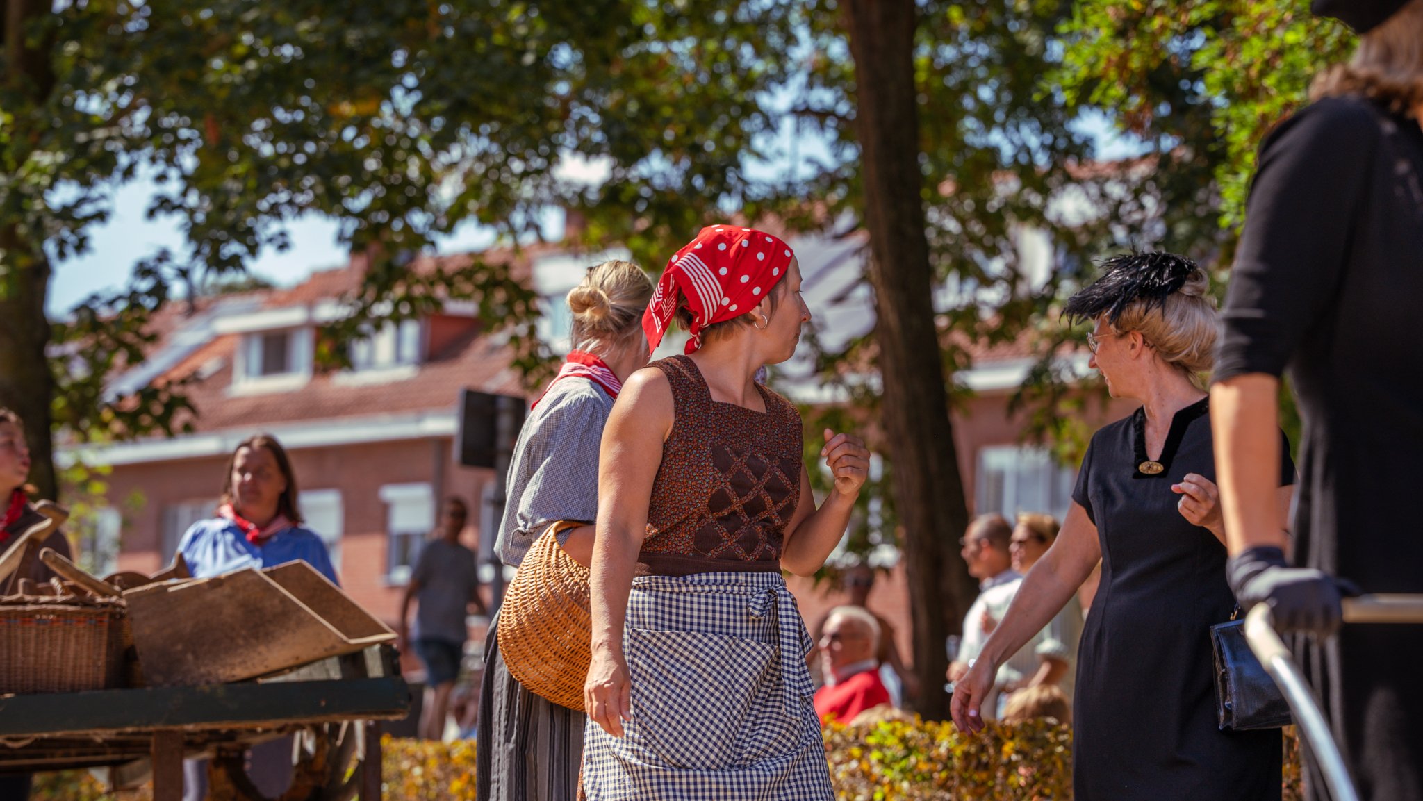 Women in vintage clothing and headscarves at an outdoor market or festival during fall, with trees and buildings in the background.