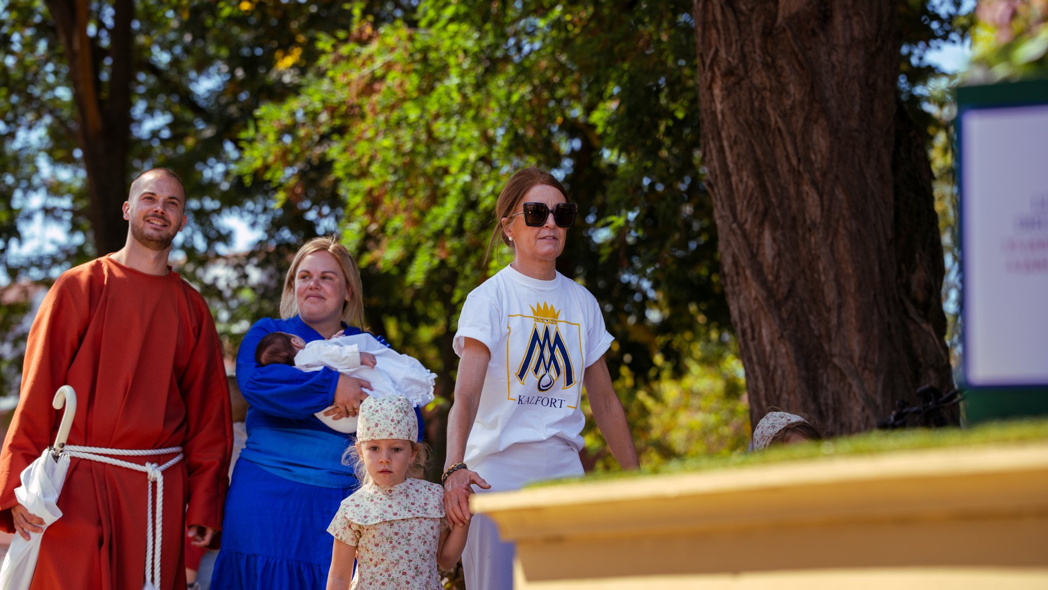Group of people outdoors with a large tree in the background, including a man in a red robe, woman holding a baby, woman in sunglasses wearing a white t-shirt with a logo, and a young girl holding hands with the woman.