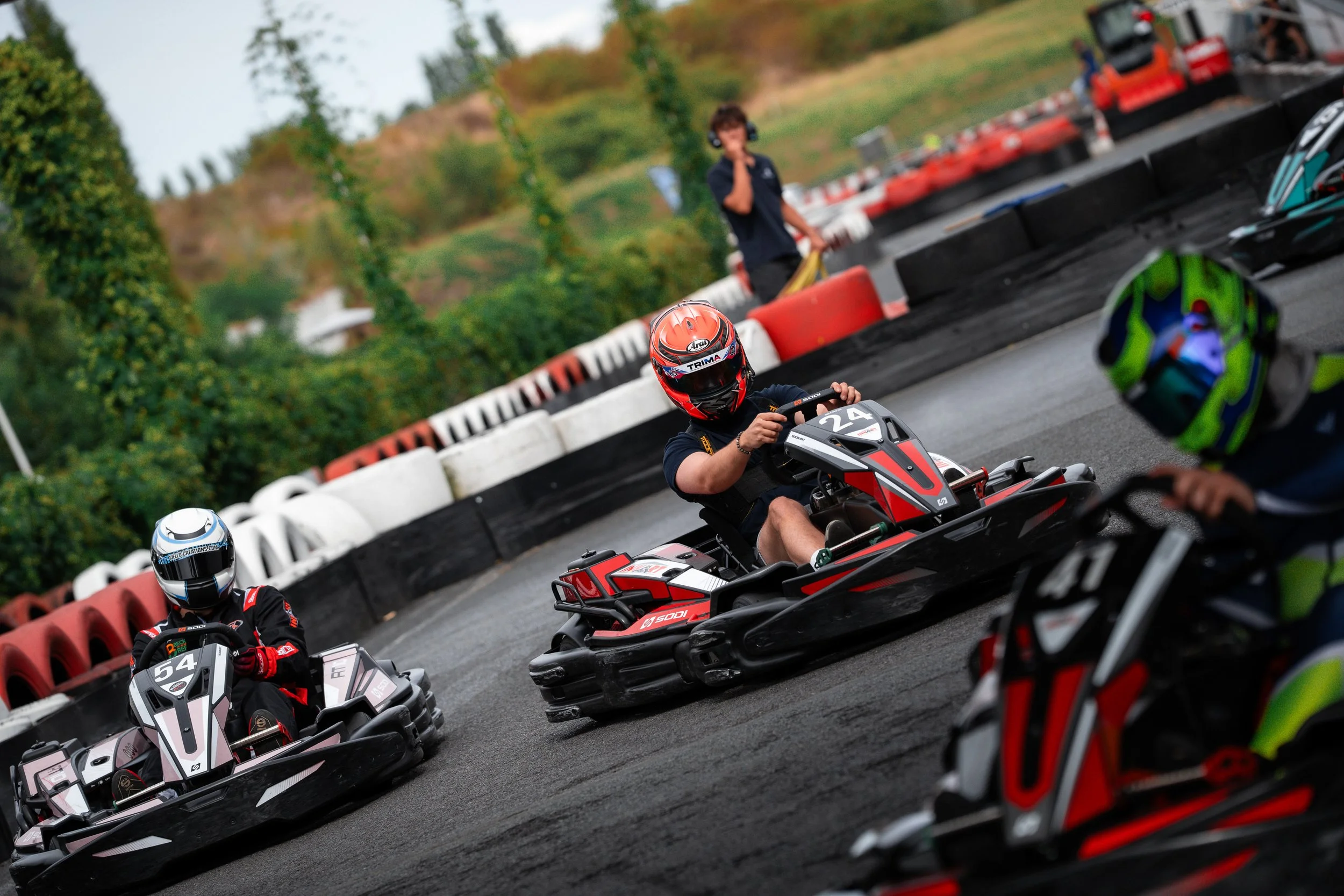 People riding and preparing go-karts on a race track surrounded by safety barriers and lush green trees.