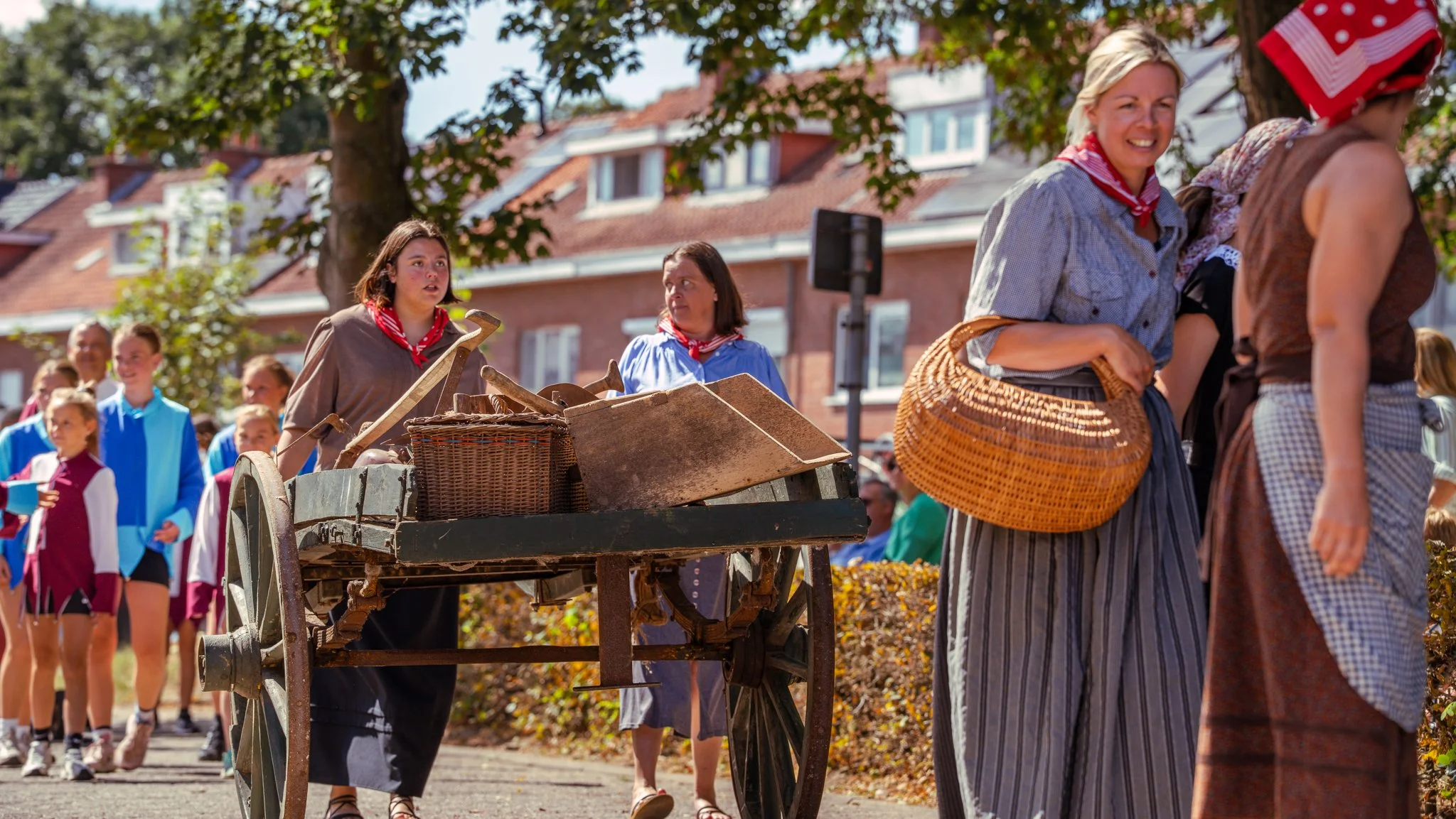 People participating in a historical reenactment parade outdoors on a sunny day, with women wearing vintage costumes and carrying baskets and carts, and children dressed in period clothing.