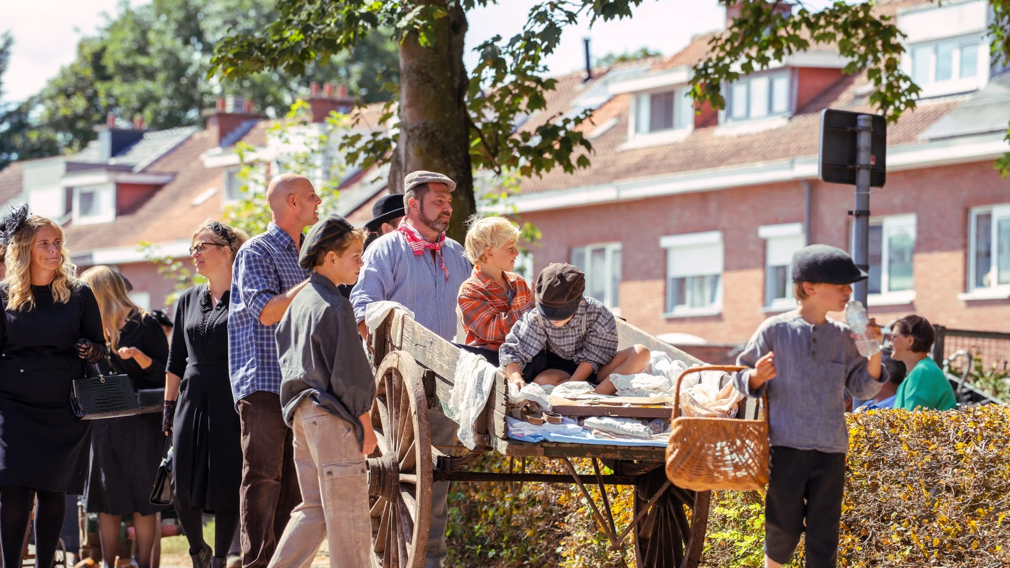 People dressed in vintage clothing at a historical reenactment, gathered around an old-fashioned wooden cart with fabric and textiles on display, in a sunny outdoor setting with trees and brick buildings in the background.