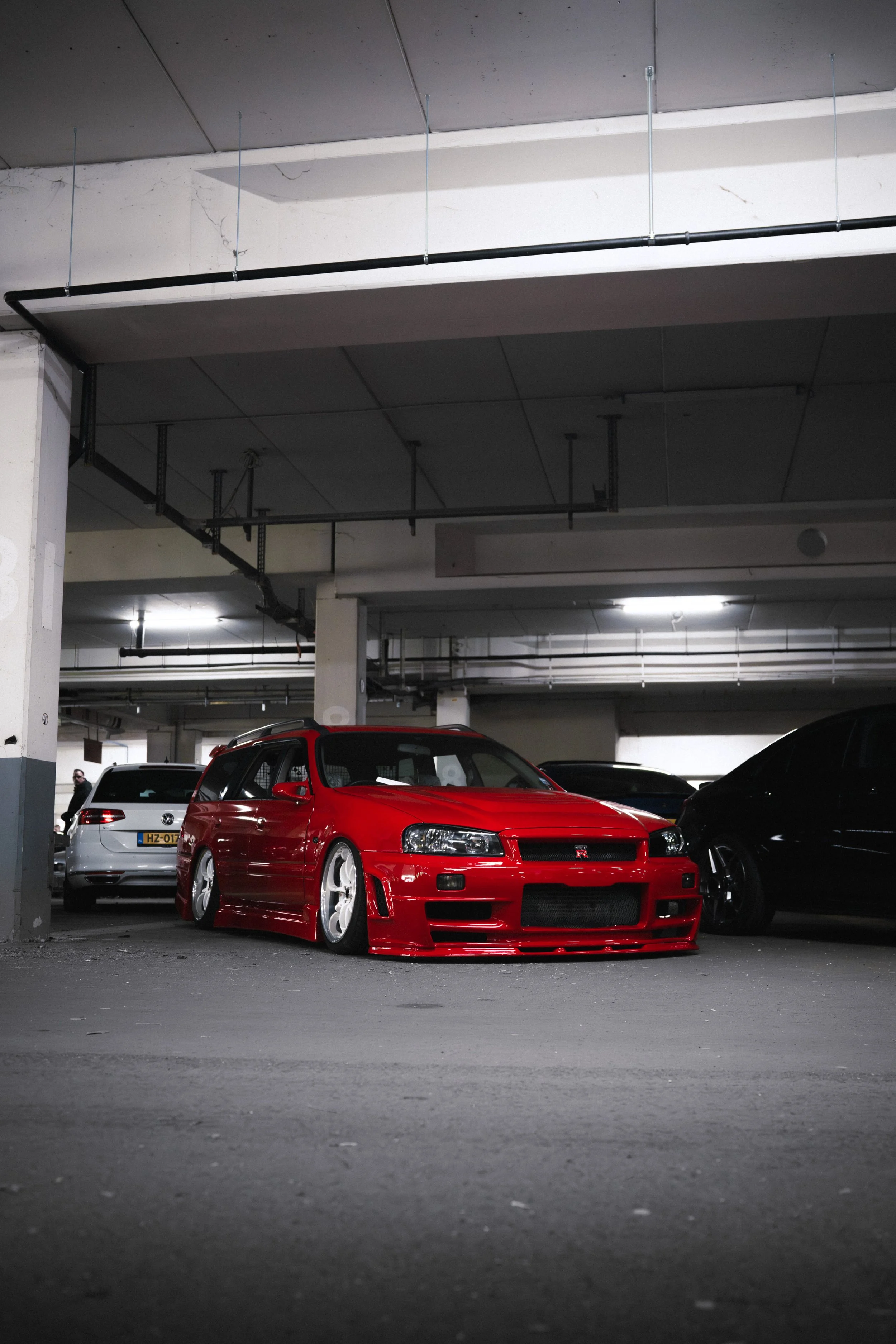 Red sports car parked in an underground parking garage next to black and white cars. The garage has concrete columns, ceiling, and visible pipes.