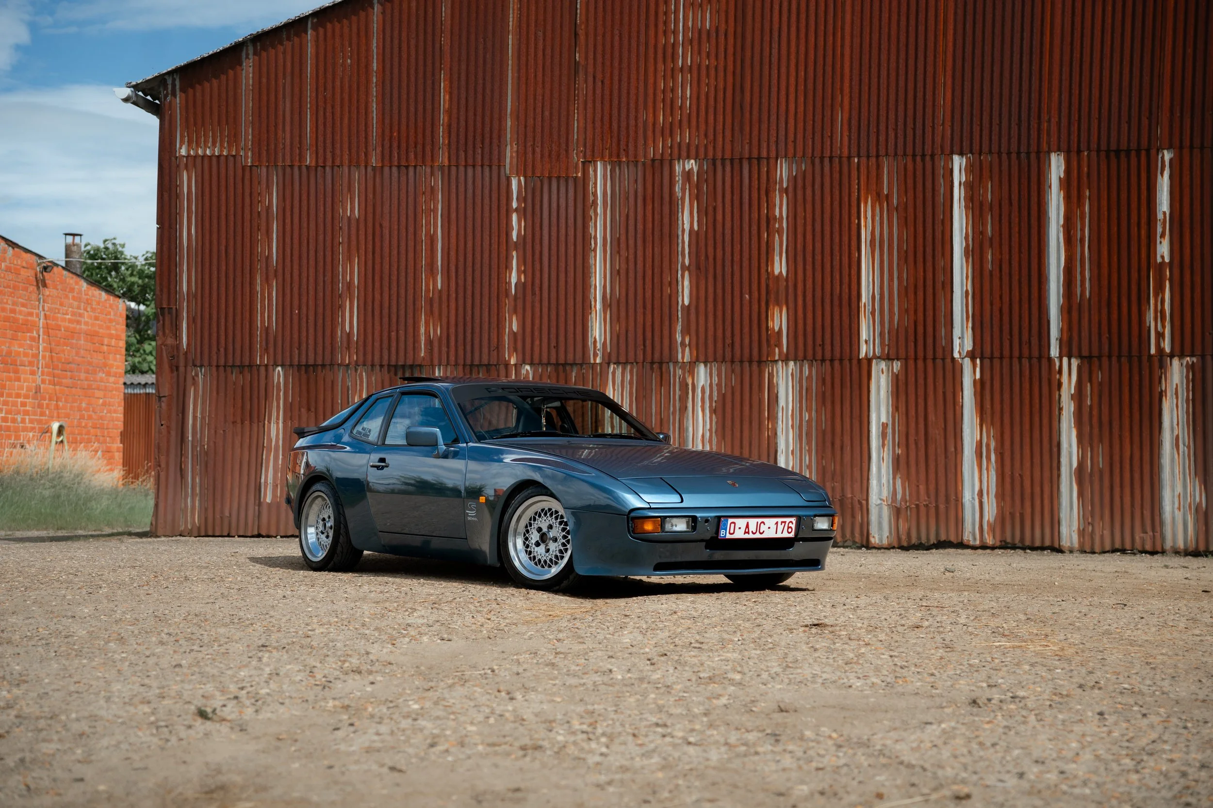 A classic gray Porsche sports car parked in front of a large weathered red metal building with a corrugated surface, on a gravel ground under a partly cloudy sky.