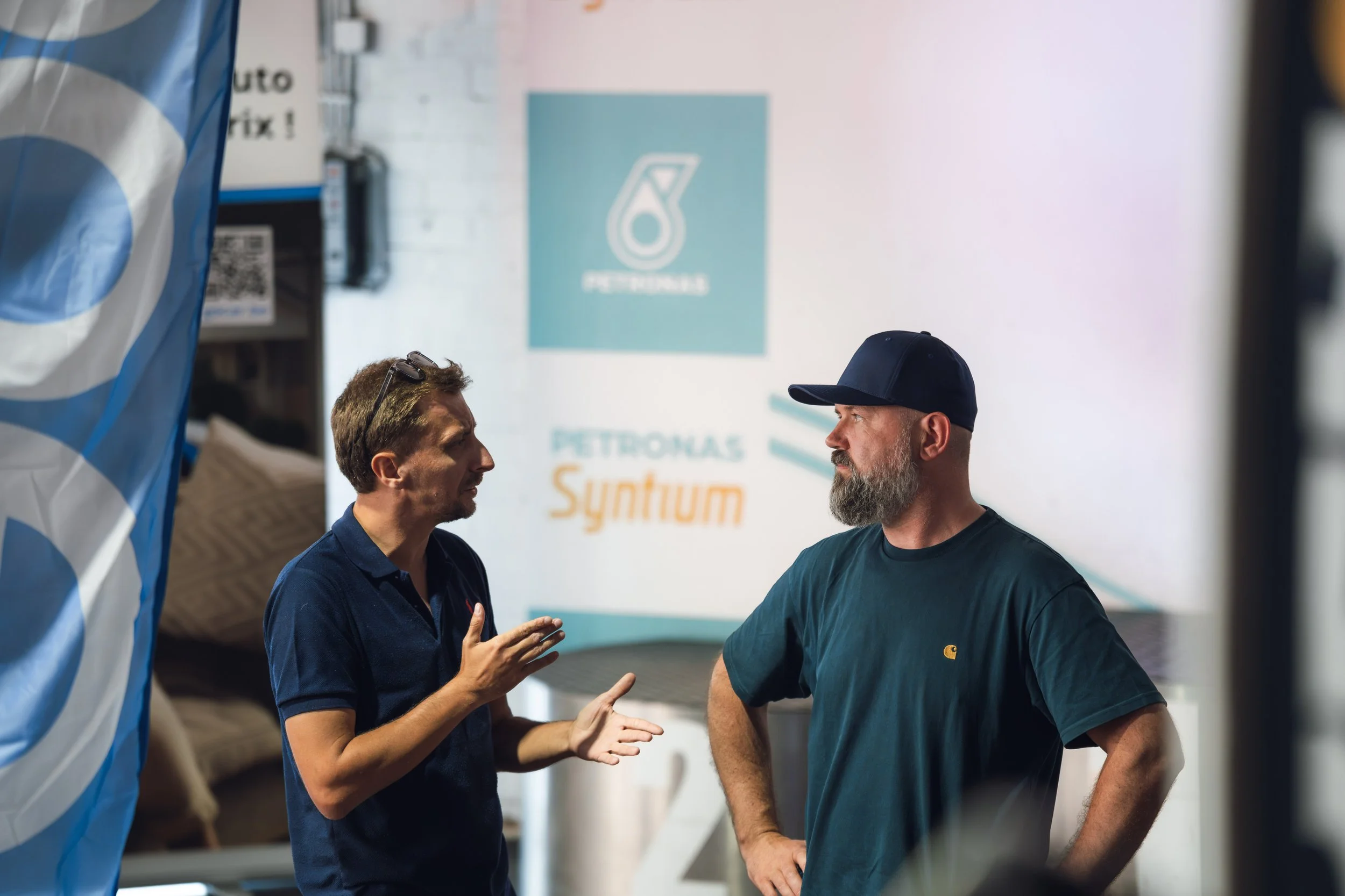 Two men are engaged in a conversation at an indoor event, with a PETROGAS sign in the background. One man, with short light brown hair and wearing a navy polo shirt, appears to be explaining something to the other man, who has a beard, mustache, and 