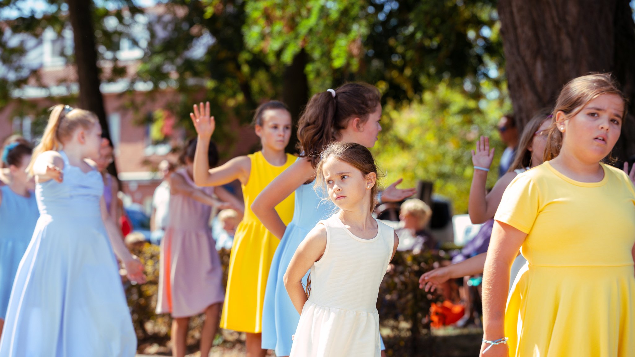 A group of young girls dressed in colorful dresses, standing outdoors in a park with trees, some with their hands raised and others with serious or confused expressions.