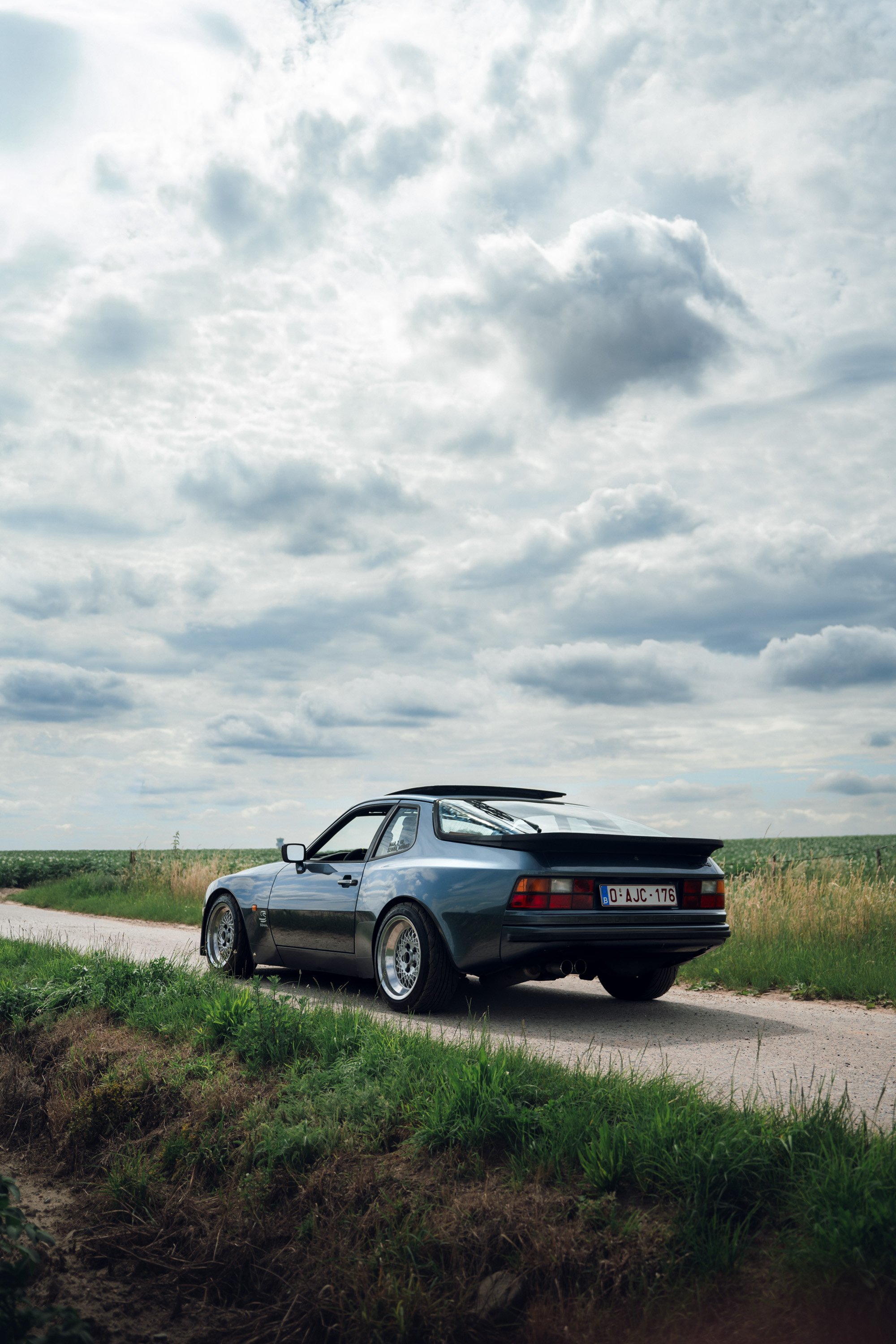A black vintage sports car parked on a dirt road in a rural area with green fields and cloudy sky in the background.