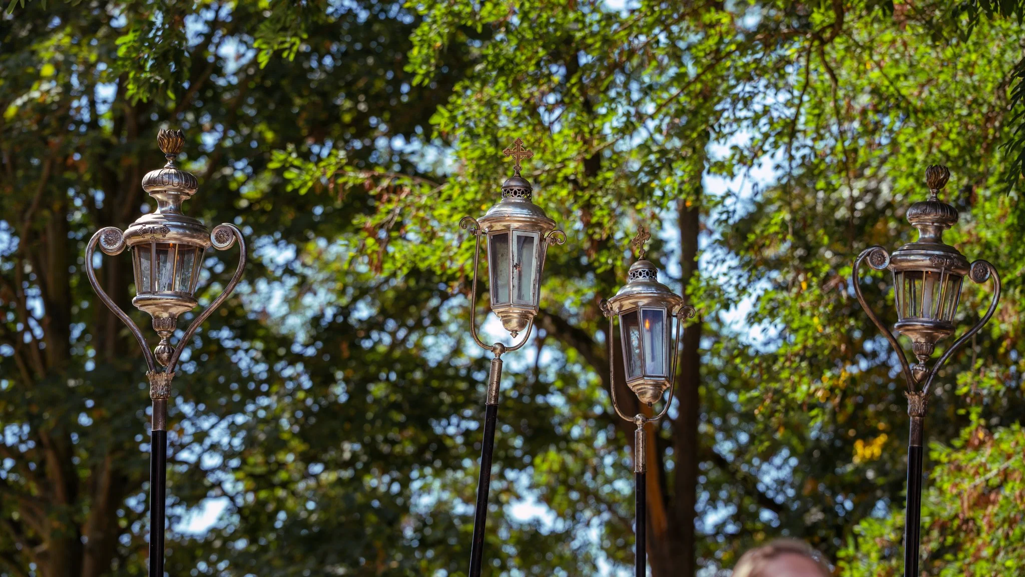 Four decorative vintage street lamps with glass enclosures and ornate metalwork, standing outdoors against a backdrop of green trees and blue sky.