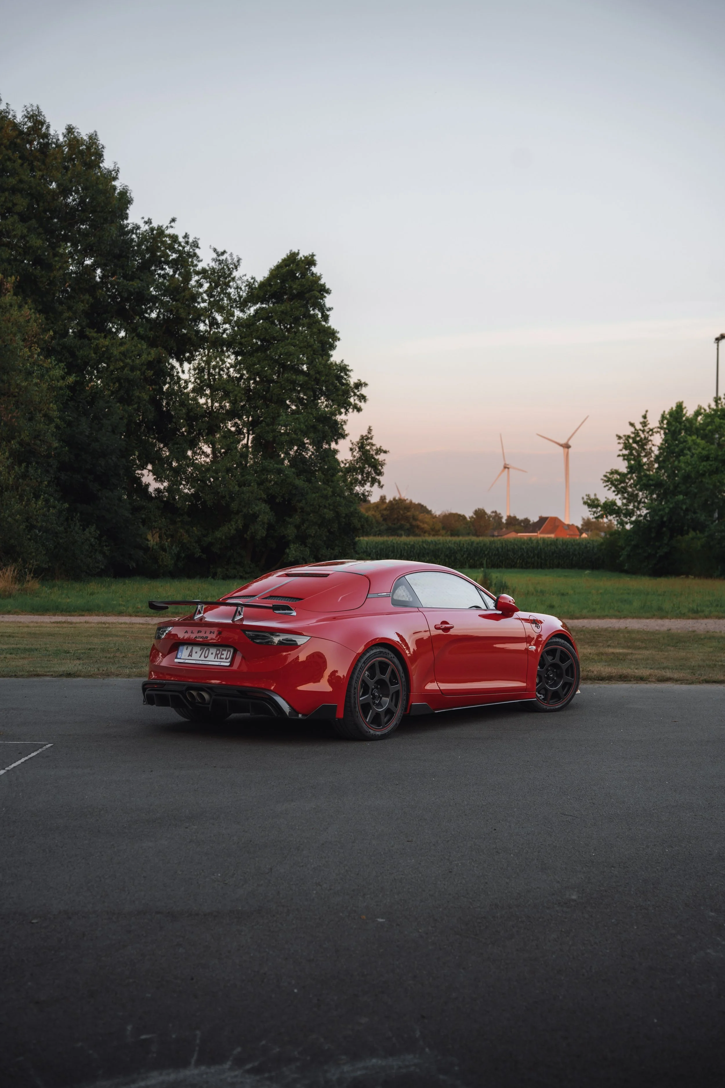 Red sports car parked on a paved lot with trees and wind turbines in the background during dusk.