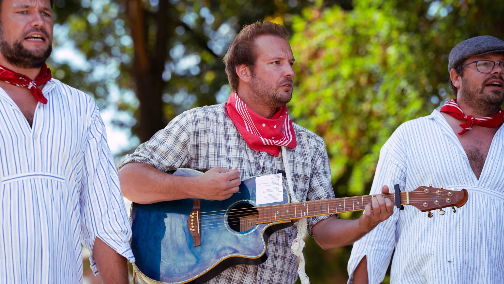Three men dressed in traditional country-style clothing, with red bandanas around their necks, standing outdoors with trees in the background. The man in the middle is playing a blue acoustic guitar.