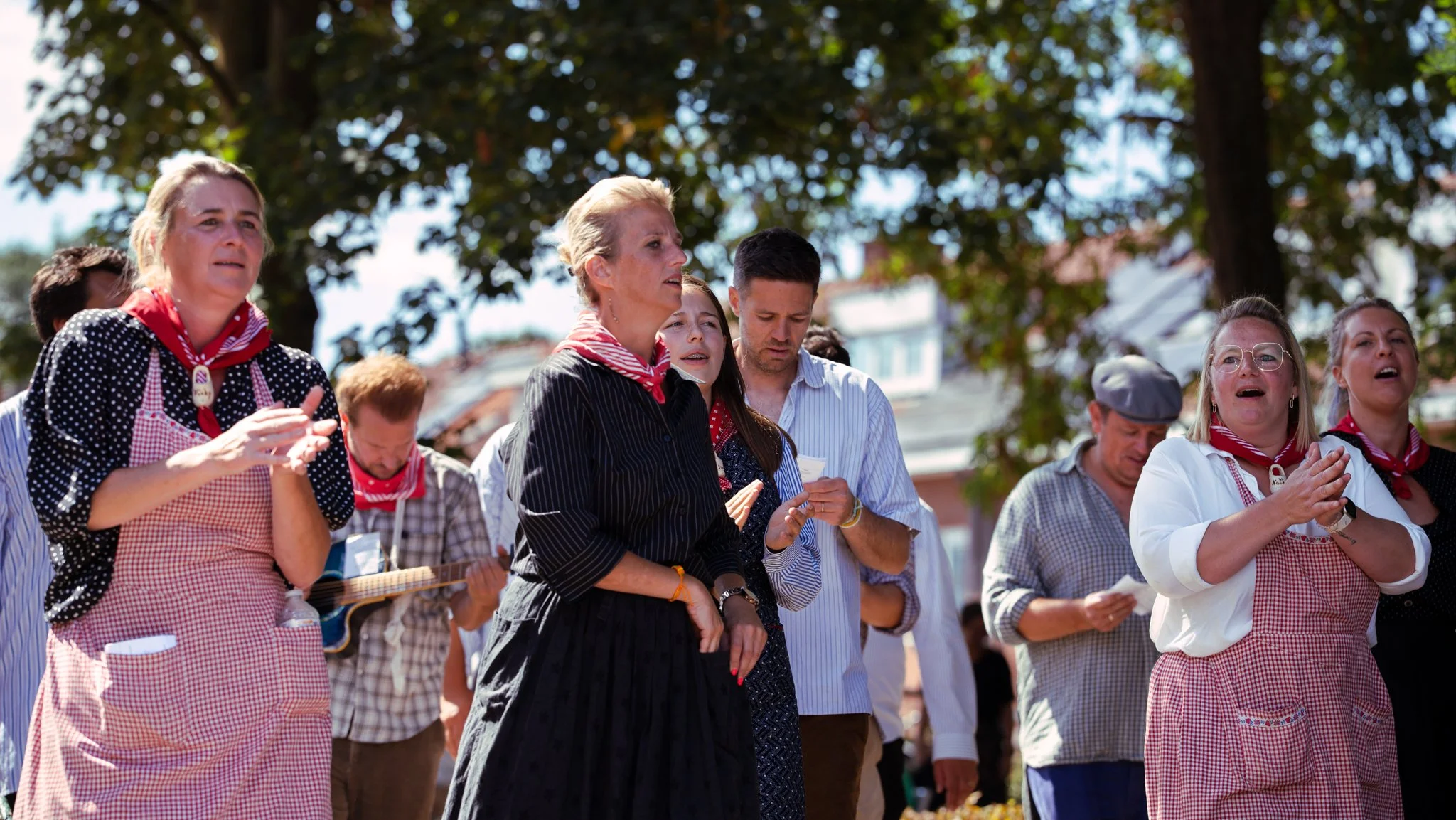 Group of people outdoors, some with hands clasped, participating in a community event under trees, wearing casual and traditional clothing, with some in red bandanas.