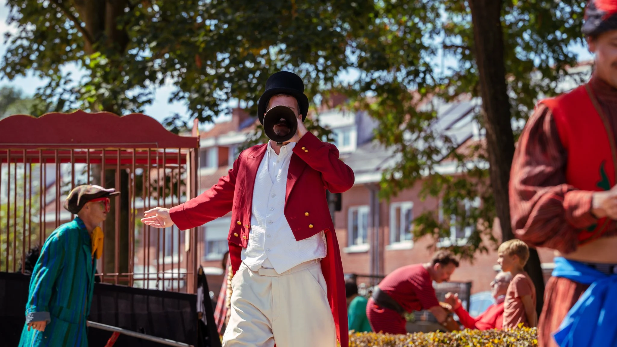 A street performer dressed as a circus ringmaster with a red coat, white shirt, and black top hat, speaking into a megaphone during a daytime parade or festival. Other festival attendees in colorful costumes are visible in the background.