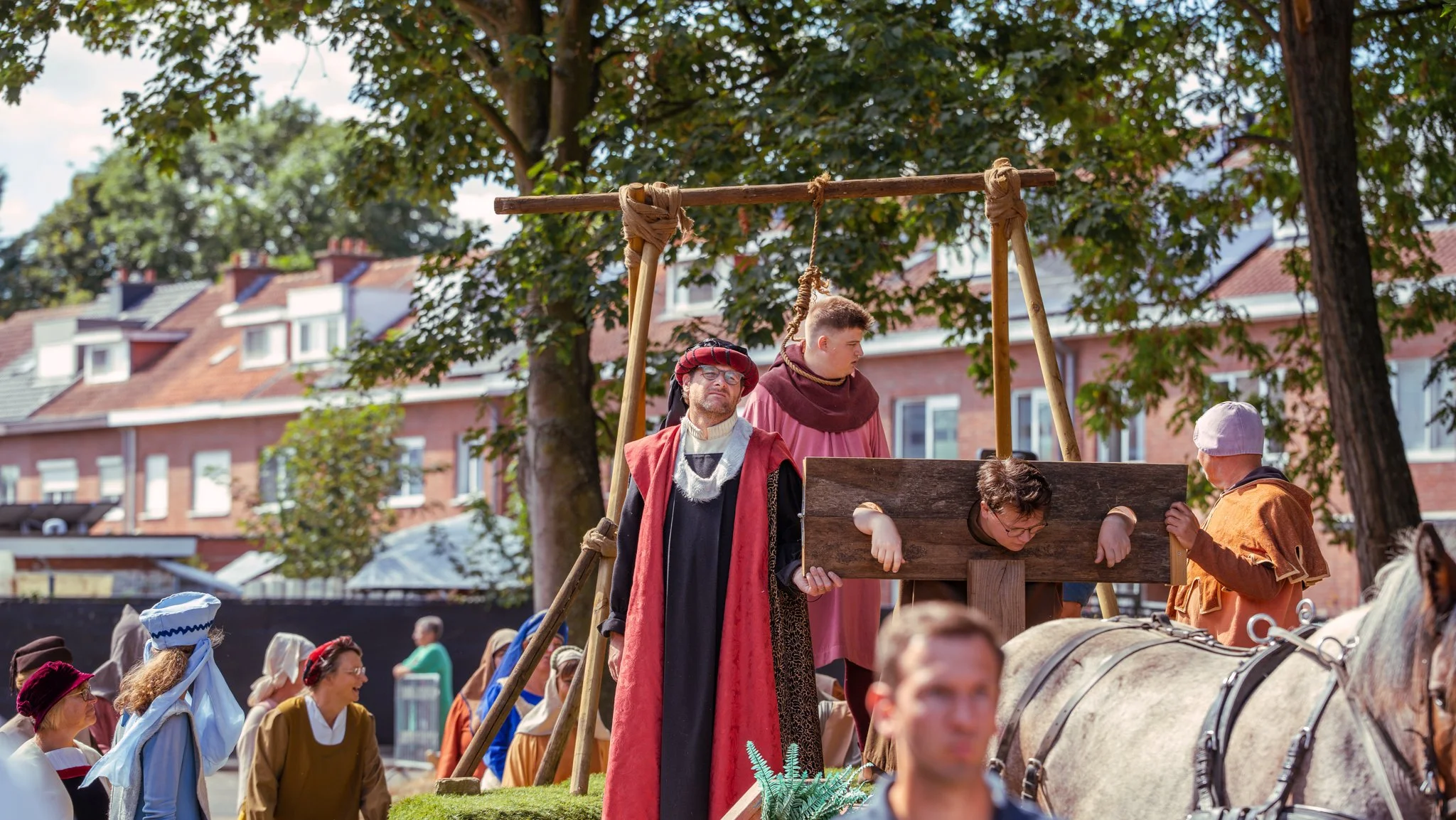 People in historical costumes reenacting a scene with a wooden rack and a person tied up, outdoors with trees and buildings in the background.