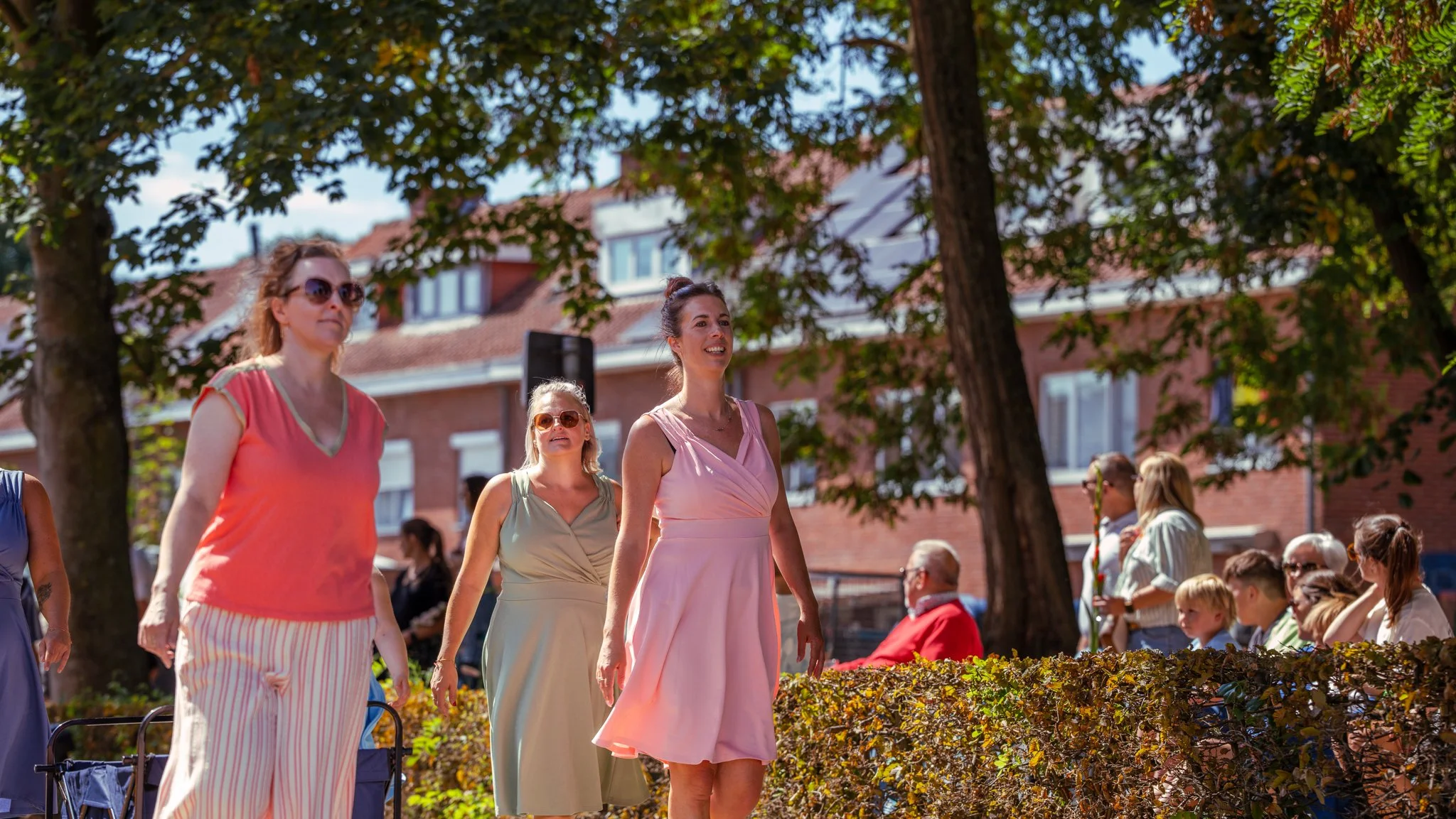 Women walking outdoors in a park during a sunny day, with trees and a brick building in the background. Spectators seated on benches are visible to the right.