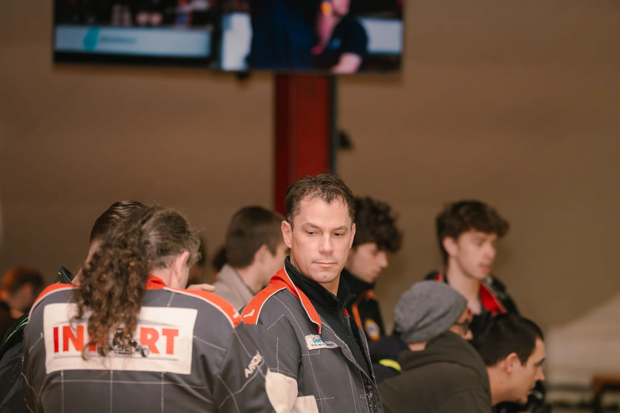 A group of men seated at a table wearing racing jackets, with one man in focus looking down and appearing contemplative, against a background of a urethane wall and a television screen.