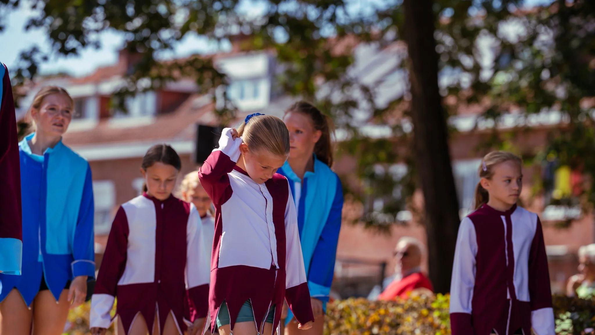 Group of young girls and women in colorful athletic costumes standing outdoors, with trees and buildings in the background.