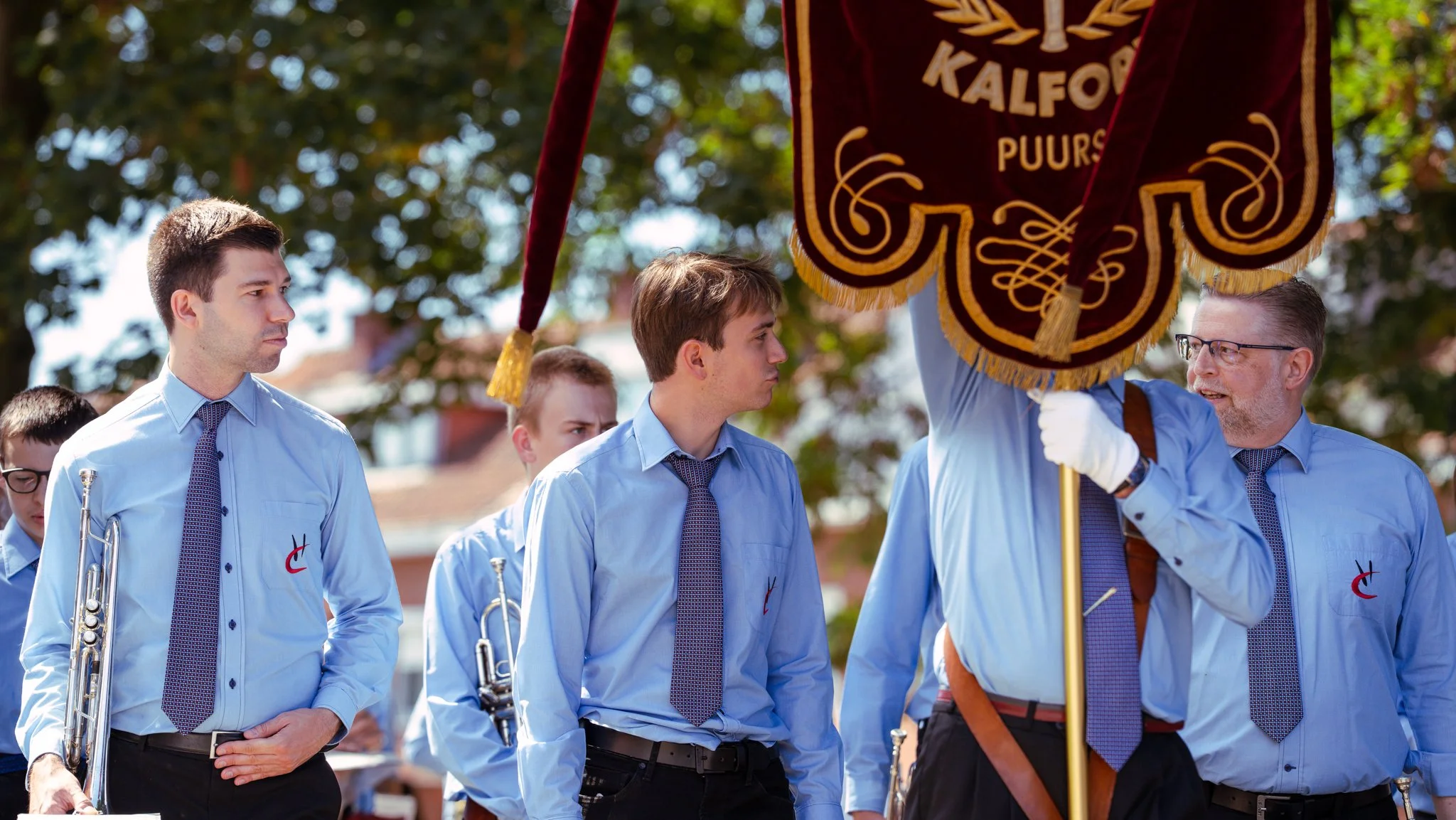 A group of men in blue shirts and ties, some holding musical instruments, stand outdoors. One man on the right holds a large ceremonial banner with the word "KALFOR" and additional text. Trees and sunny weather are visible in the background.