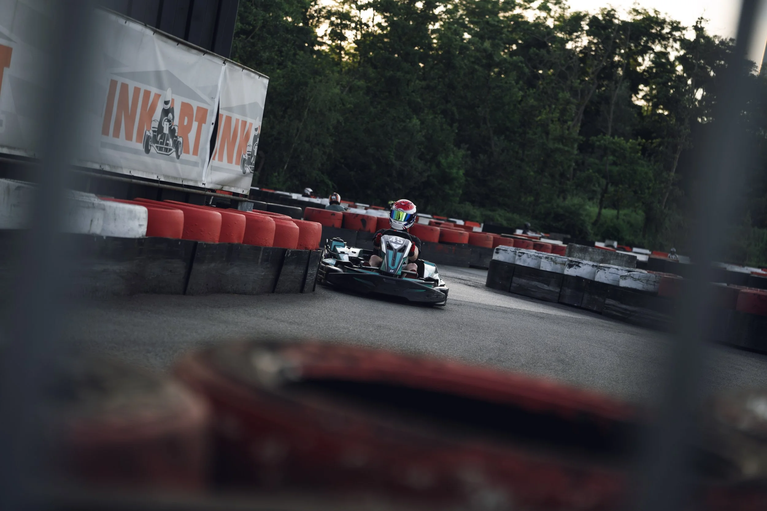 A person racing in a go-kart on an outdoor track surrounded by trees, wearing a helmet and racing gear, with orange and black tire barriers and a promotional sign in the background.