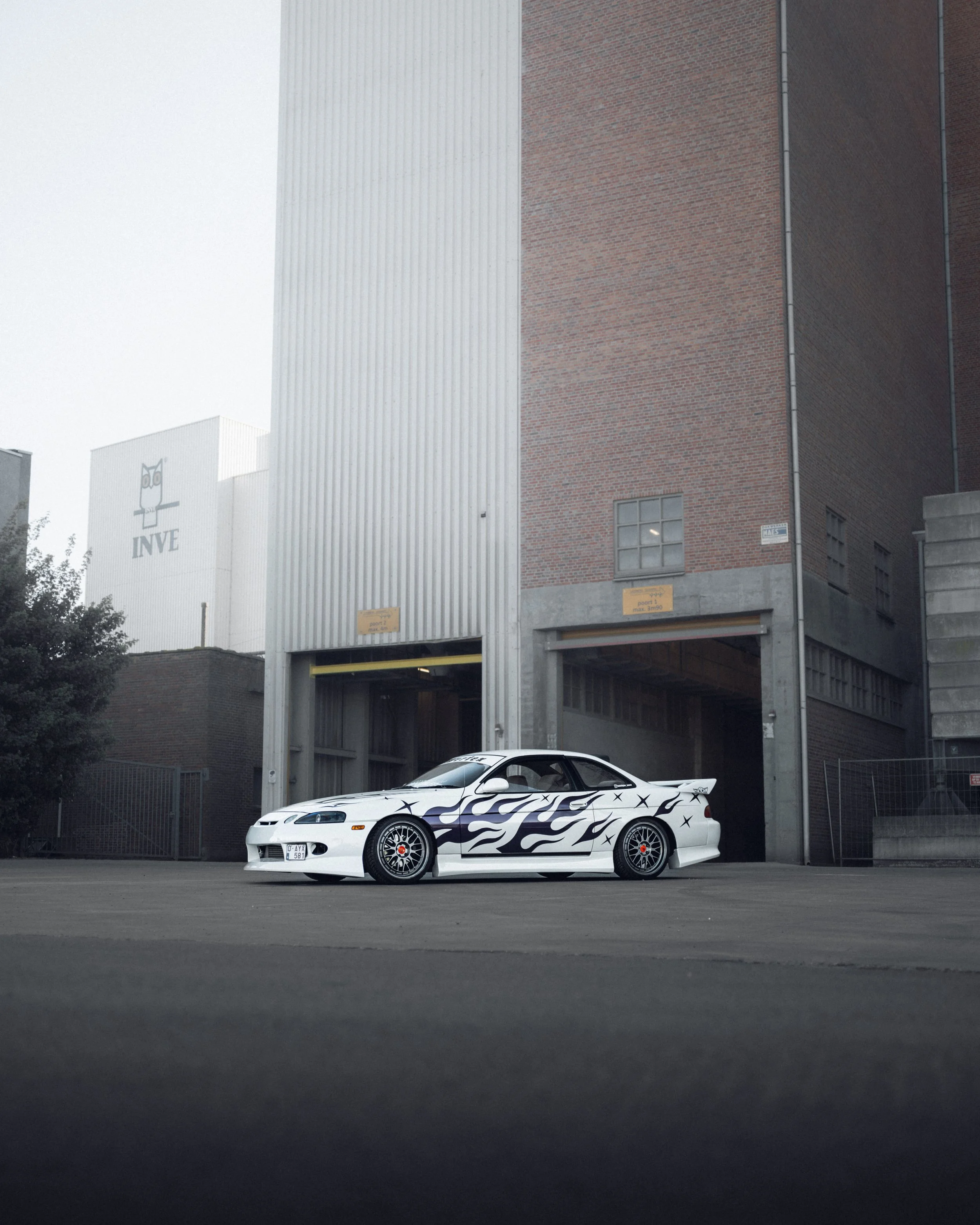 White sports car with black flames and star decals, parked in front of an industrial building.