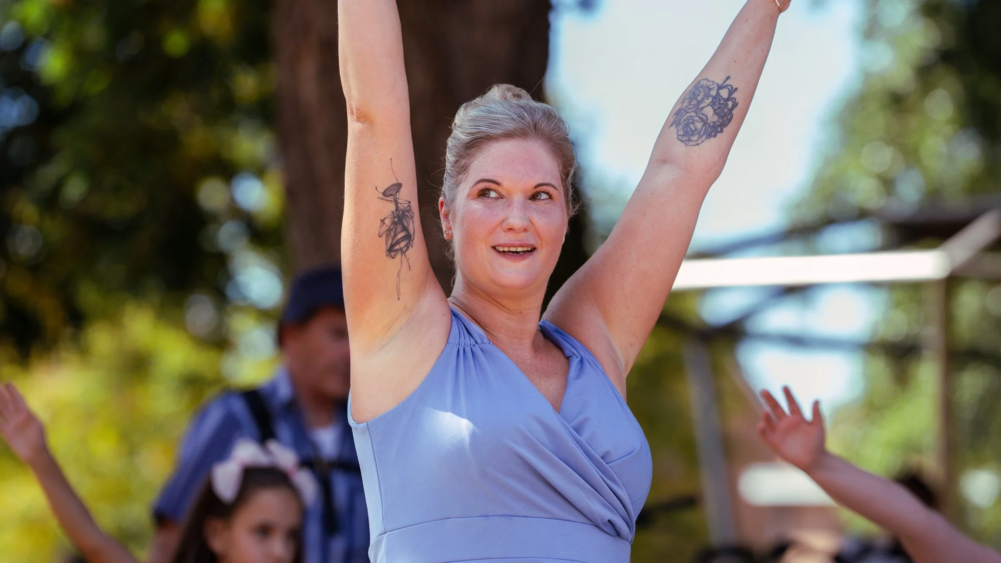 Woman with blonde hair and tattoos celebrating outdoors with arms raised, smiling, in a blue dress, with children and trees in the background.