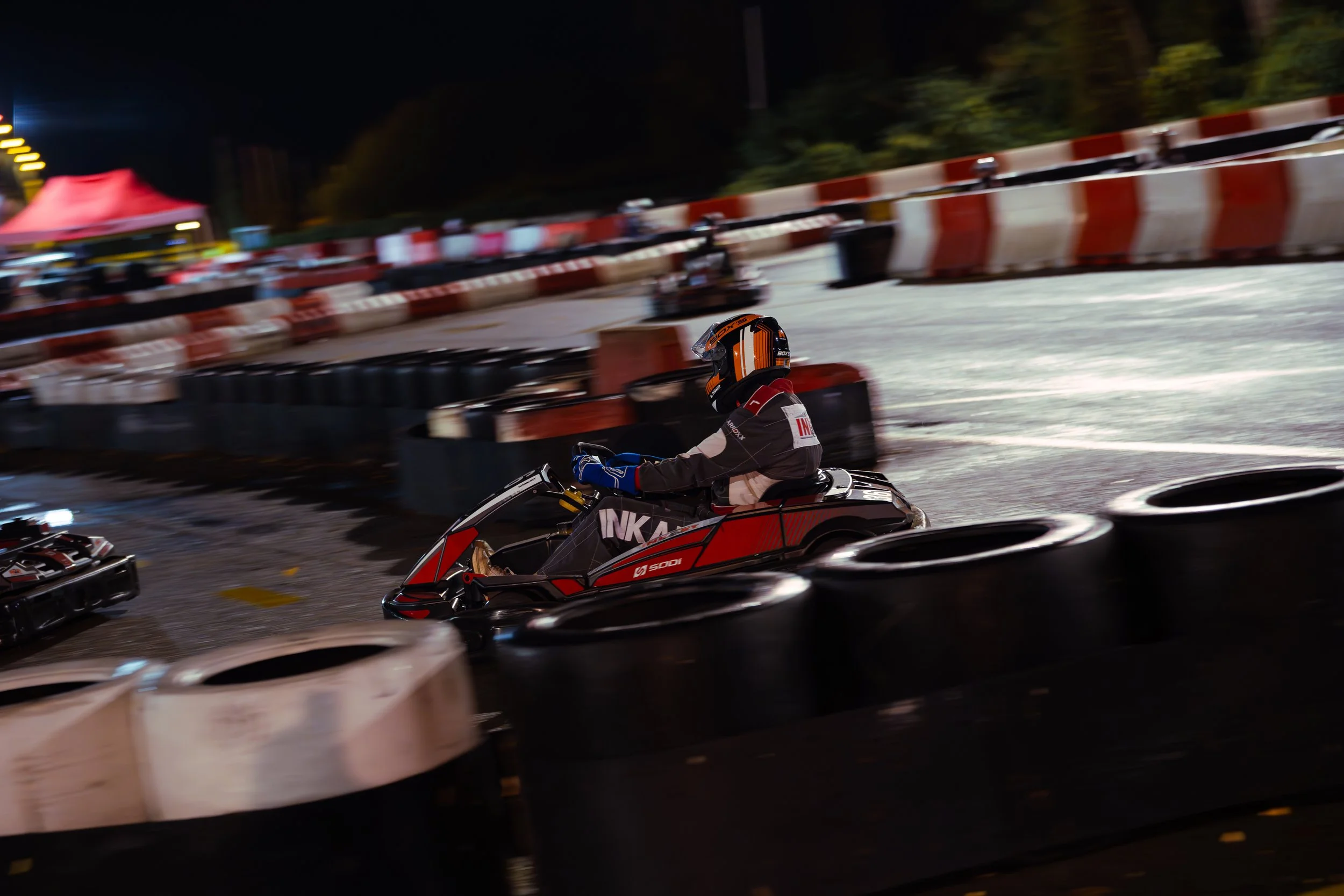 Nighttime go-kart race with a driver wearing a black and orange helmet, racing on a track secured by red and white barriers, with blurred motion indicating high speed.