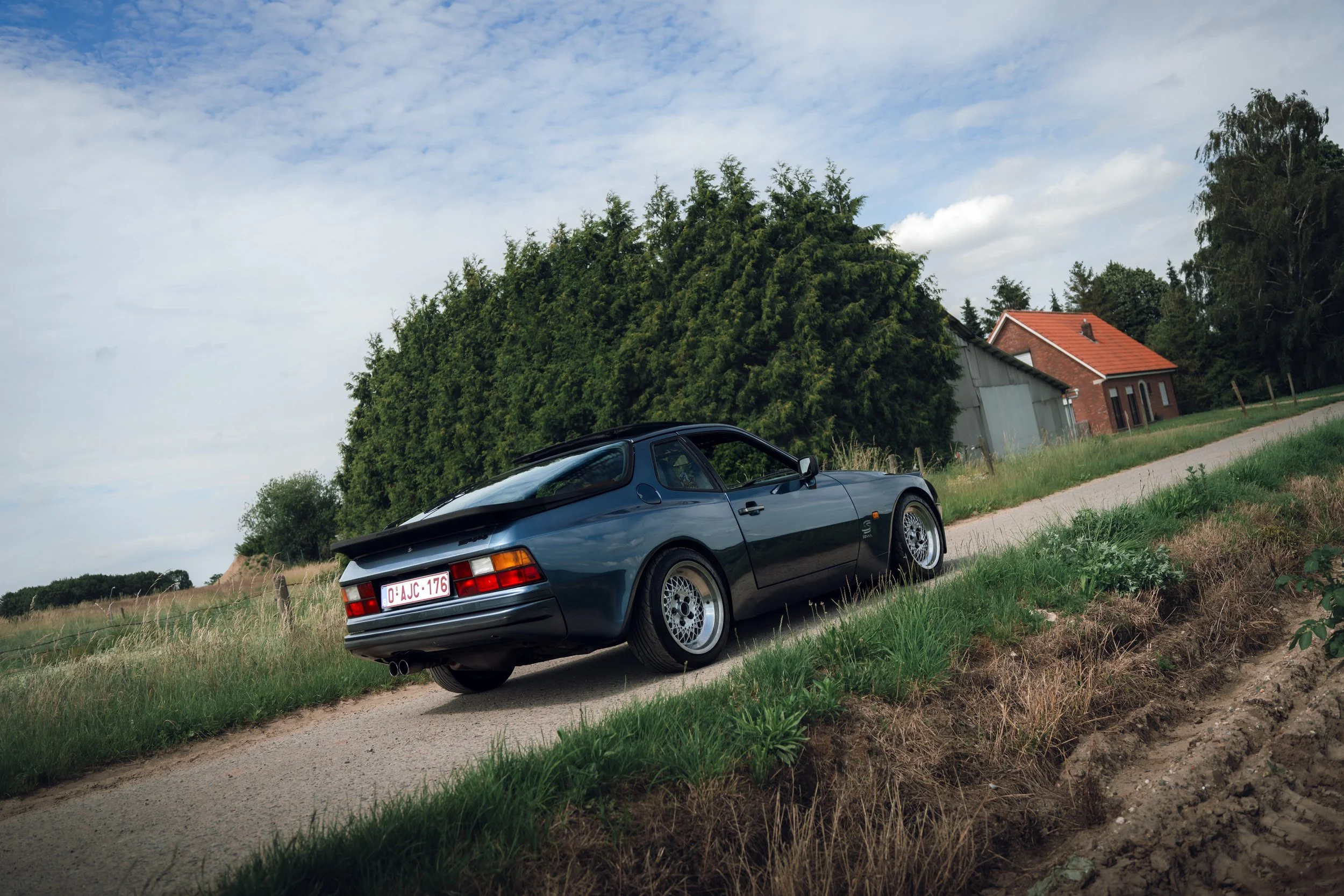 A dark gray sports car parked on a rural dirt road next to a grassy area. In the background, there's a tall green tree, a small red-brick house with a red roof, and a partly cloudy sky.