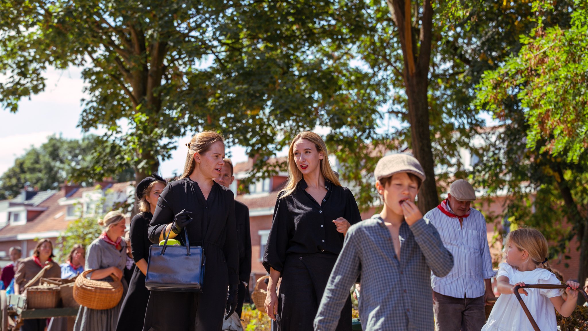 People dressed in historical clothing at an outdoor event in a park with trees and buildings in the background.