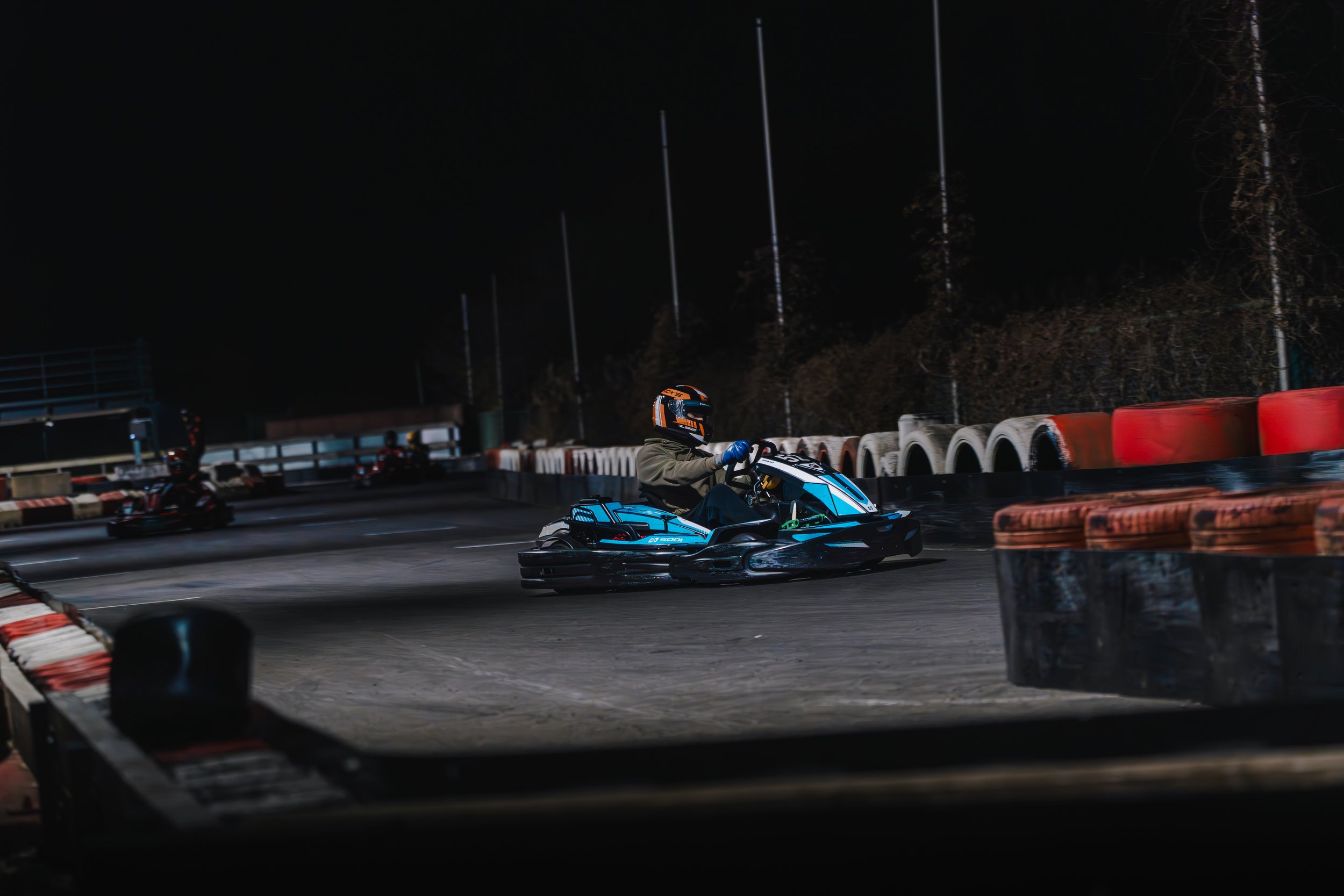 Nighttime go-kart racing scene with a driver in helmet and racing suit on a track, surrounded by tire barriers and illuminated by track lights.