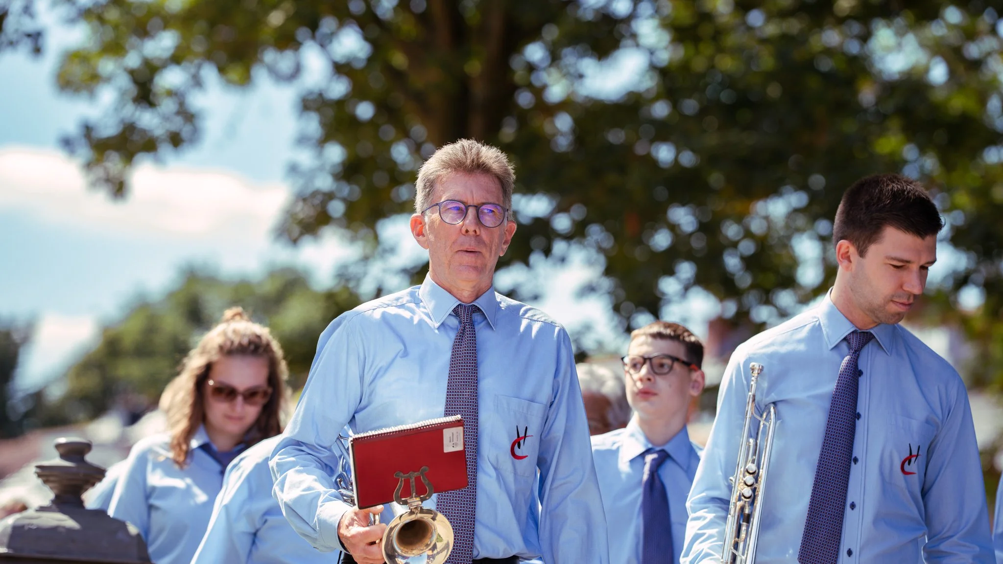 A group of four musicians walking outdoors in daylight, wearing light blue shirts and ties, with trees and blue sky in the background. The man in the center is holding a trumpet and a folder, and the others are carrying wind instruments.