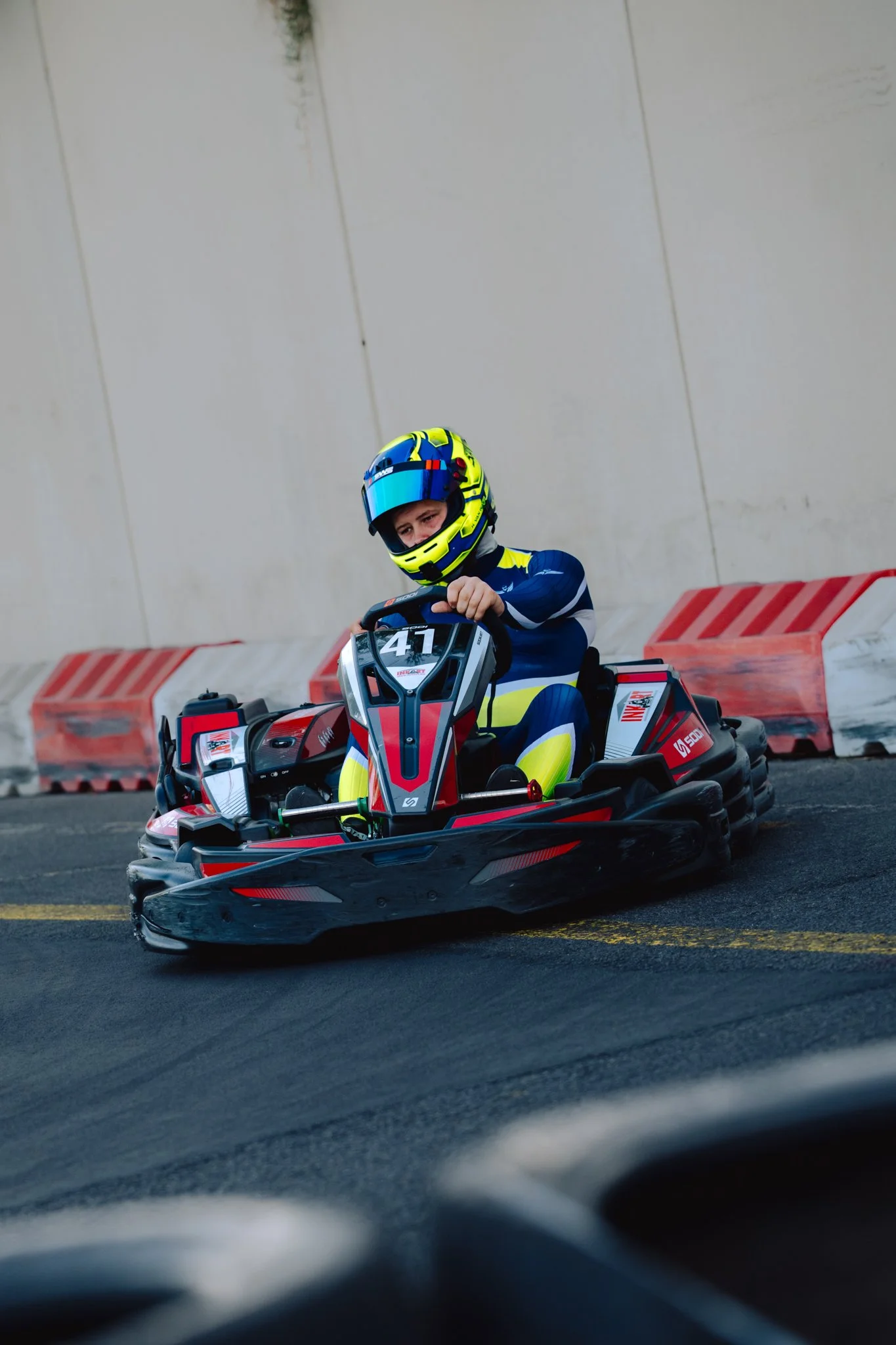 A young boy wearing a blue and yellow racing suit and a matching helmet drives a red and black go-kart on a paved track.