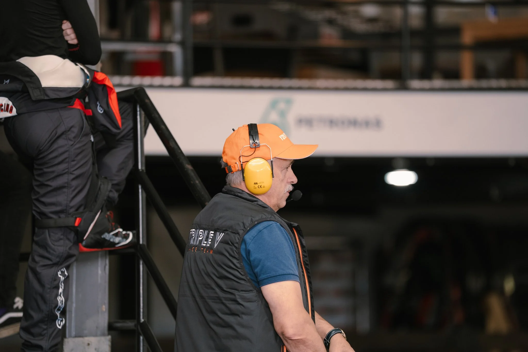 A man wearing an orange cap, black vest, and yellow noise-canceling headphones standing in a garage or pit area, watching a race or event.