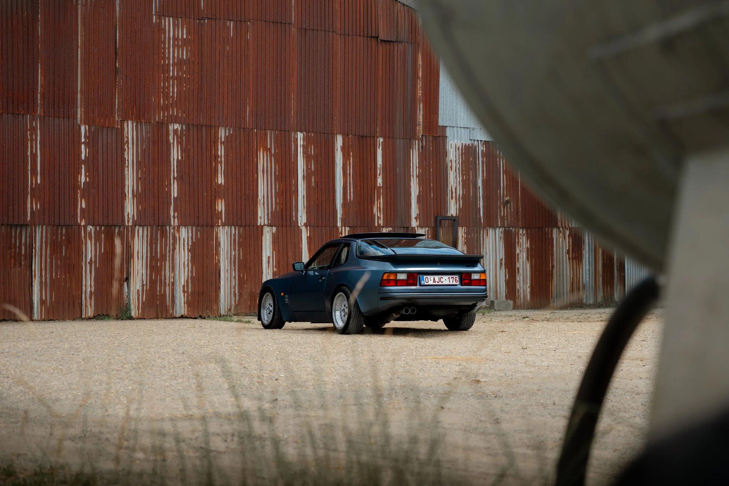 A black sports car parked in front of a large, rusted corrugated metal wall, with a partial view of a blurred object or structure in the foreground.