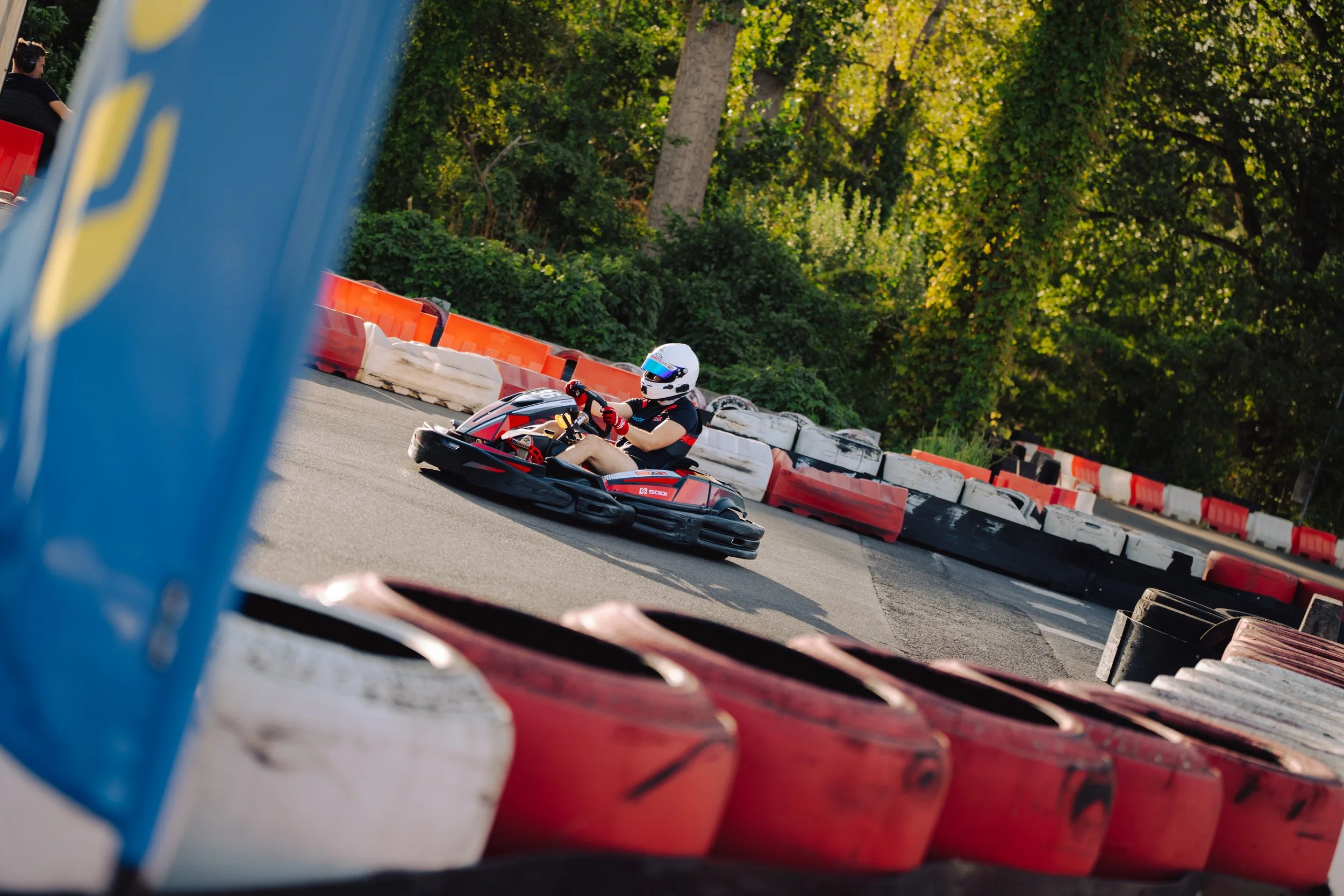 A person wearing a helmet and gloves driving a go-kart on an outdoor track with green trees in the background, partially viewed through a barrier.