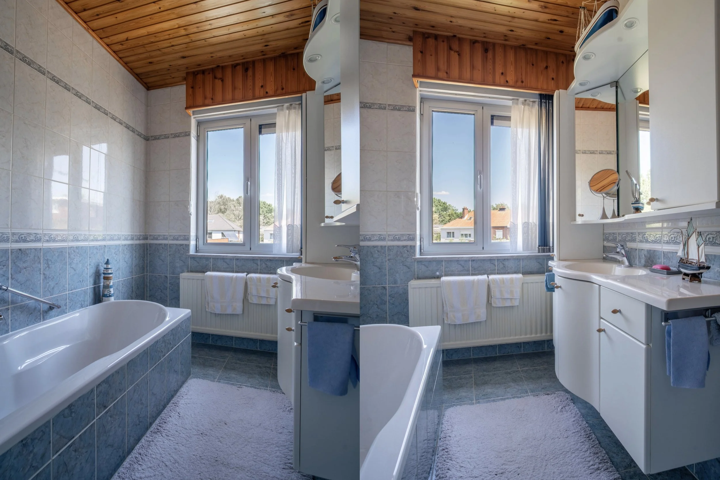 Bathroom with white and blue tiled walls, window with white curtains, wooden ceiling, mirror, sink with cabinet, bathtub, and a small lighthouse decoration.