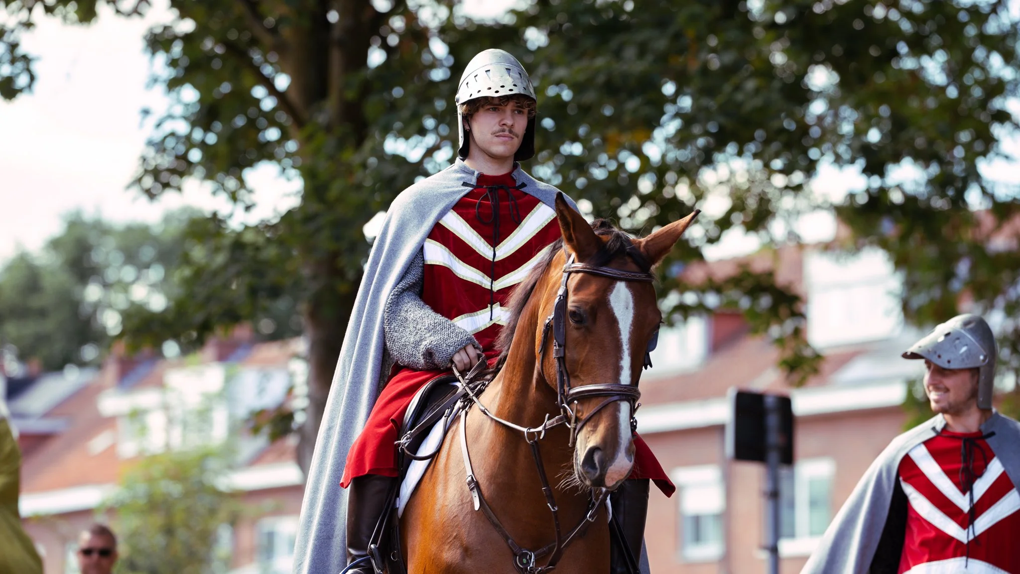 A man dressed as a medieval knight riding a horse during a reenactment event, with another person in costume standing nearby.