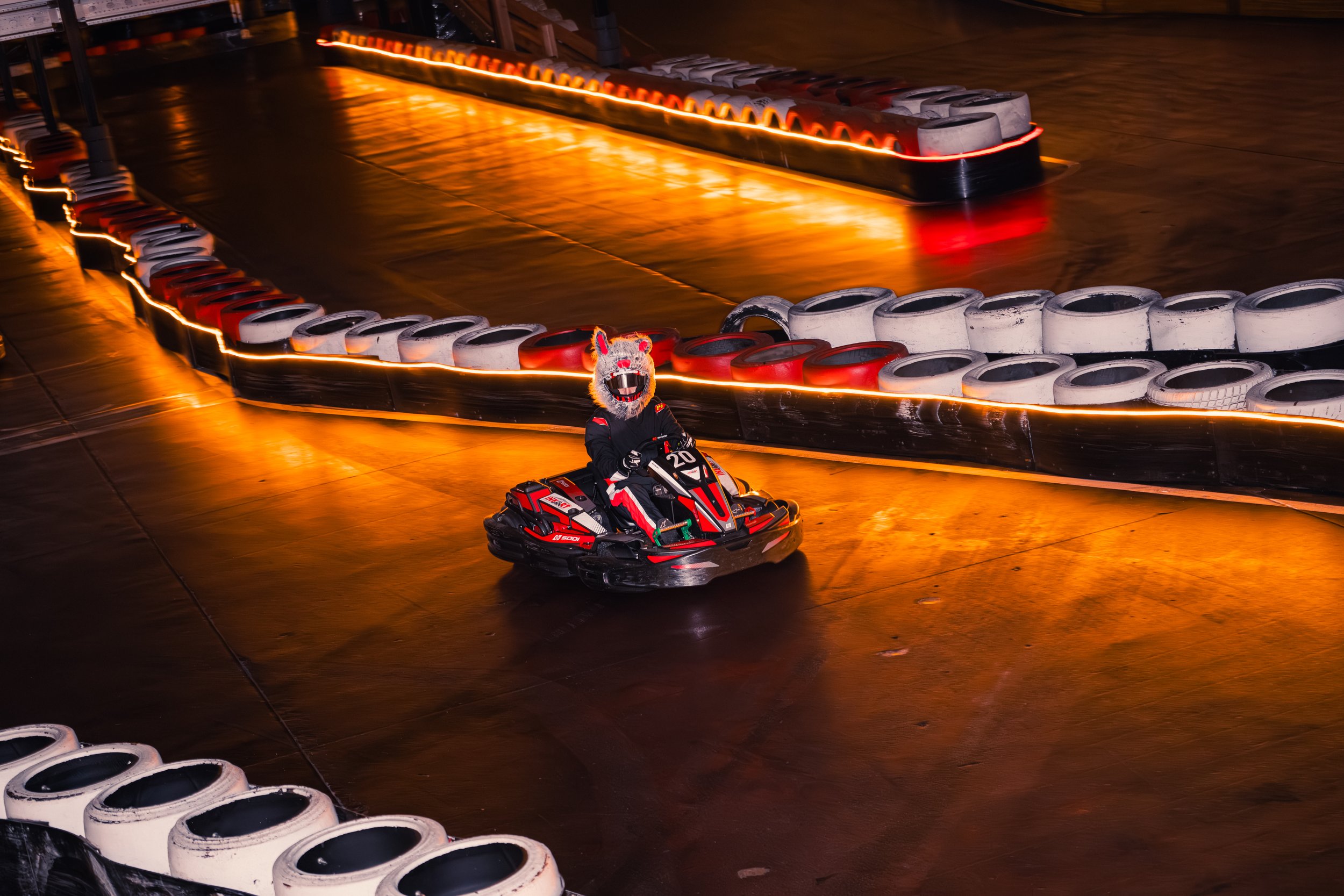 A person in a horse mask riding a go-kart on an indoor race track with illuminated barriers.