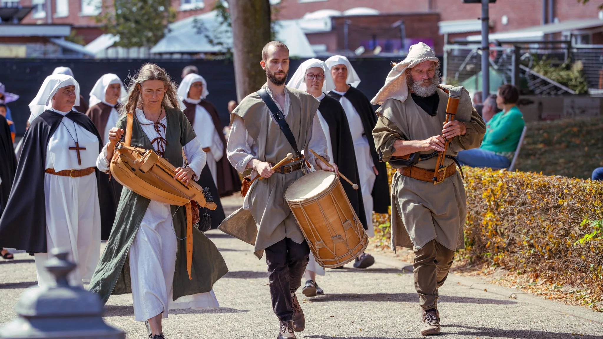 People dressed in historical clothing participating in a parade or reenactment, with some playing traditional instruments, outdoors in a park or street setting.